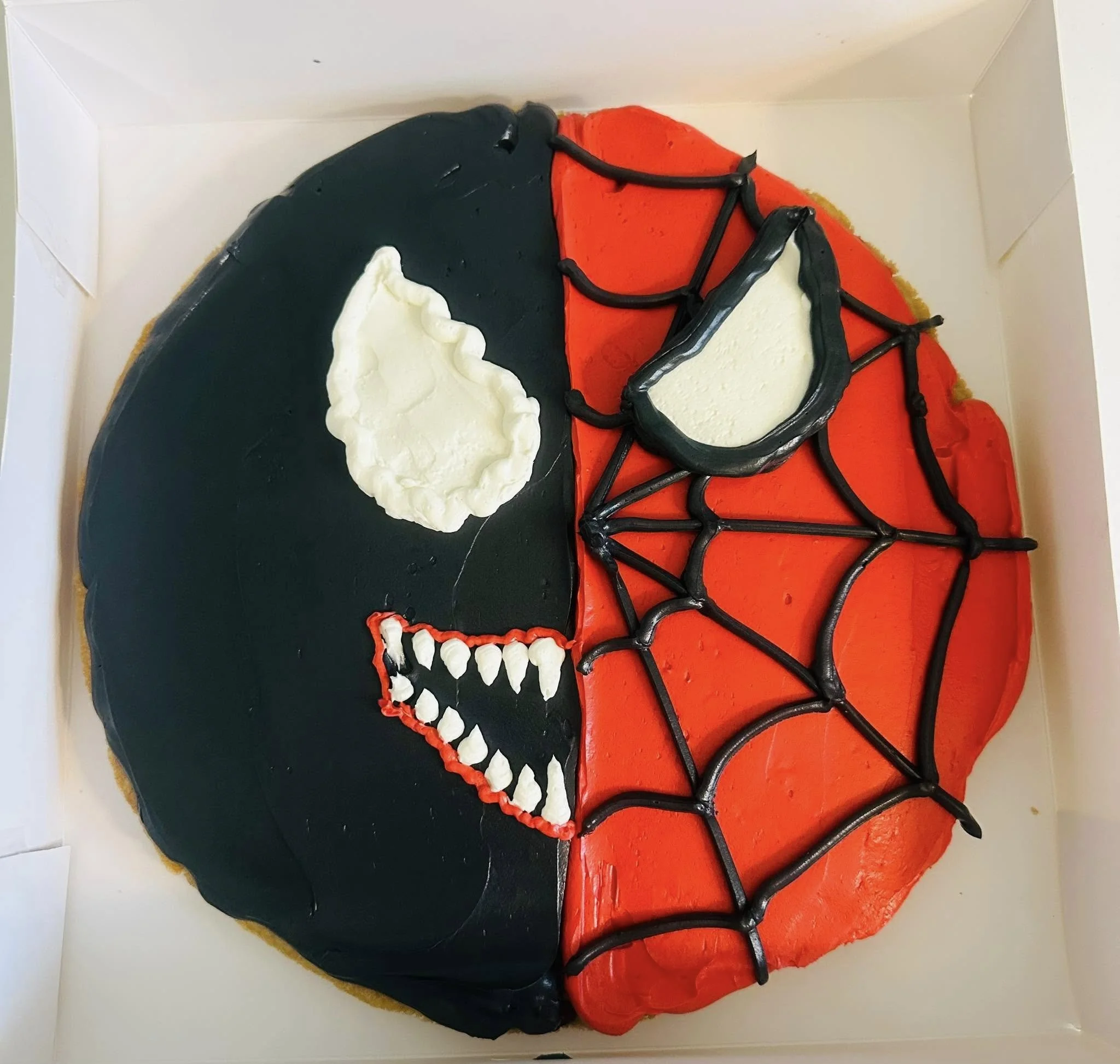 A cake decorated with a half-black, half-red design, featuring a white skull, a red mouth with sharp teeth, a white eye patch, and black web-like lines, resembling a combination of a skull and a spider web.
