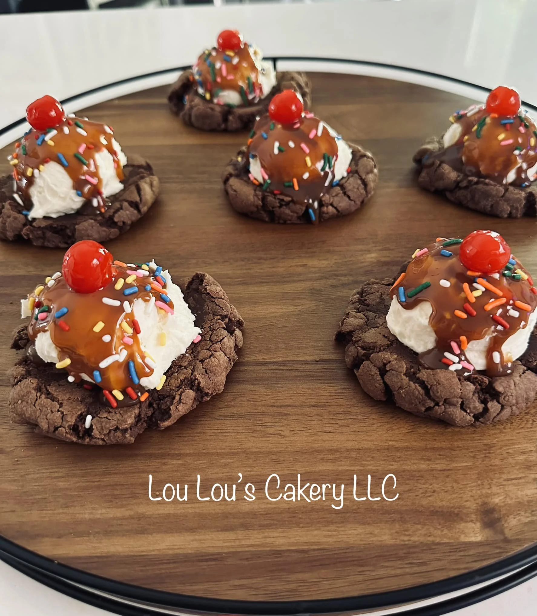 Six cookies topped with ice cream, chocolate syrup, rainbow sprinkles, and cherries on a wooden tray with the text 'Lou Lou's Cakery LLC'