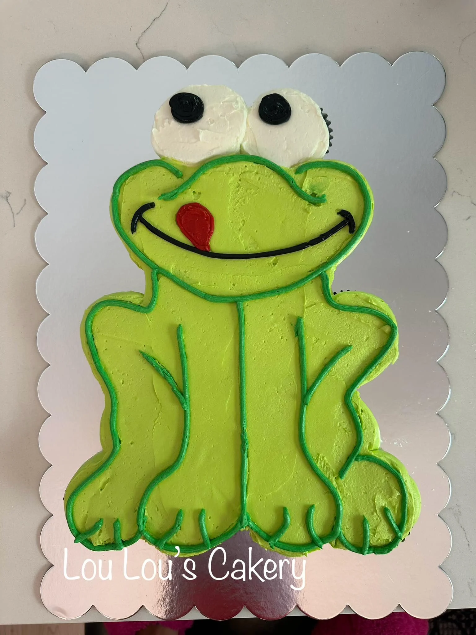 A cake decorated to look like the character Kermit the Frog from The Muppets, with green icing, black eyes, a red tongue, and a smiling mouth, on a silver scalloped cake board.