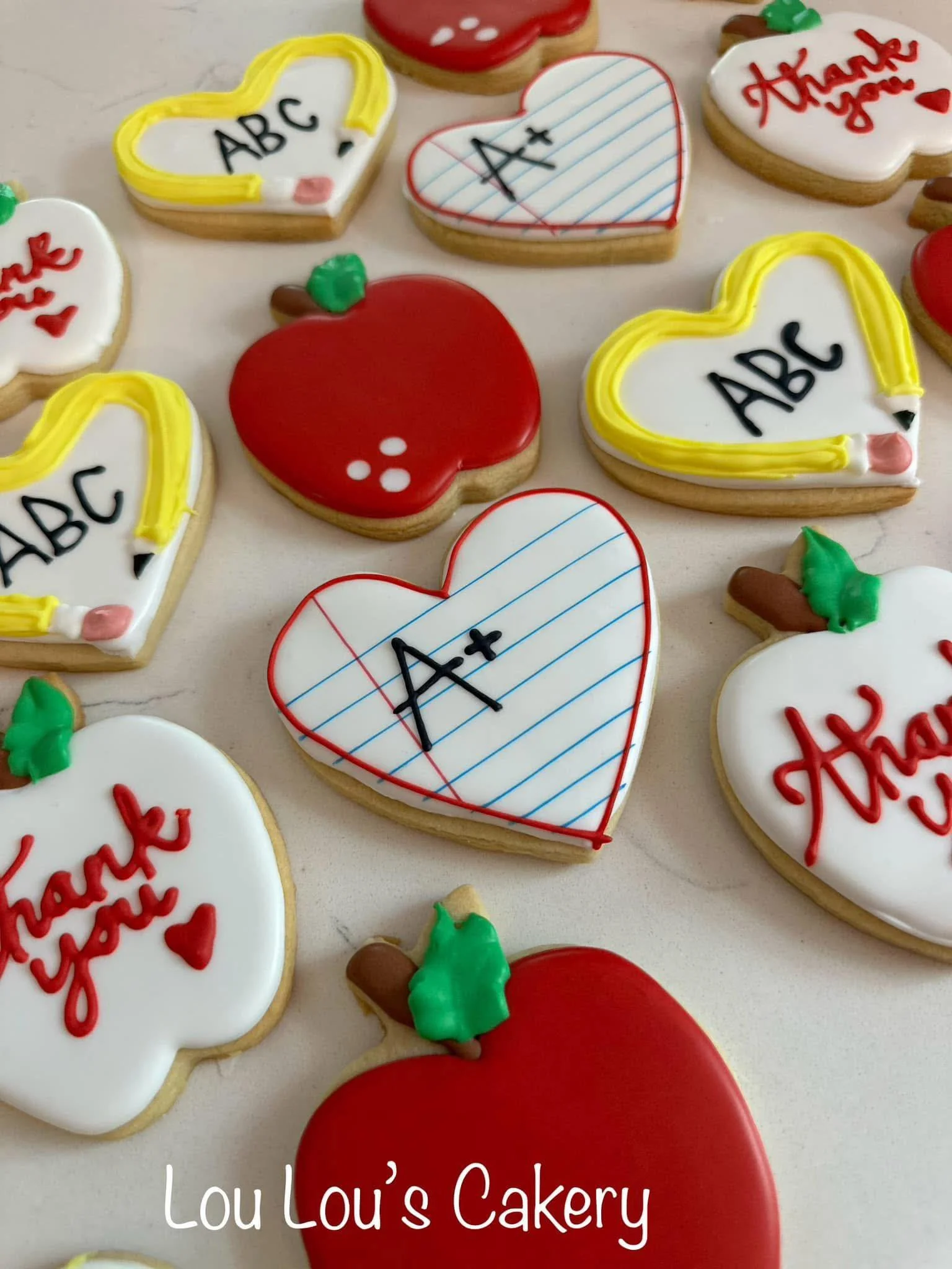 Assorted decorated cookies, including red apples, white hearts with blue-lined paper design and 'A+' grade, and white hearts with 'Thank you' written in red, on a white surface.