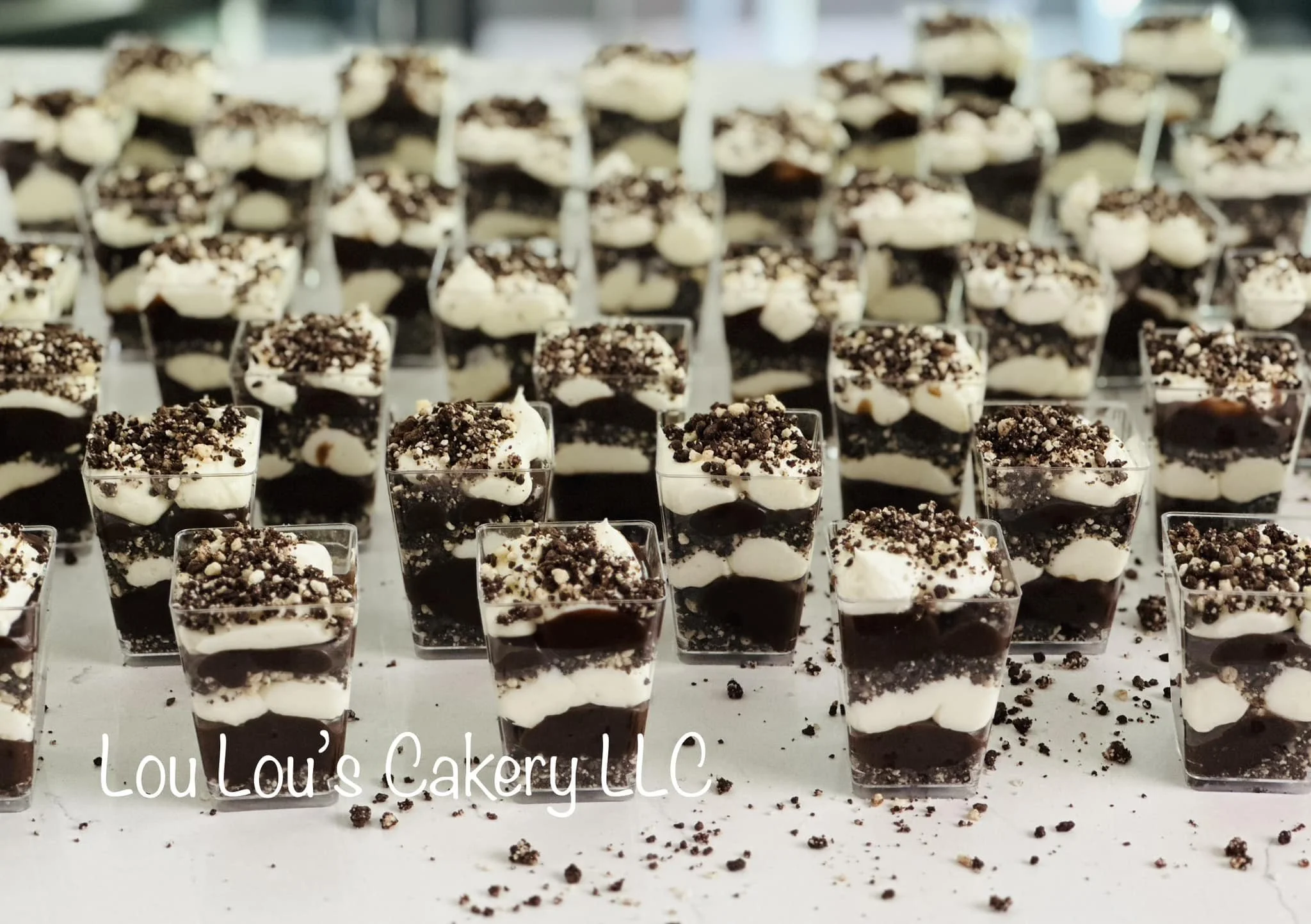 Mini layered chocolate and vanilla parfait desserts topped with crushed cookies, arranged in small clear cups on a white surface with some crumbs scattered around.
