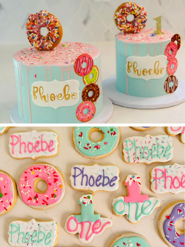 Two turquoise birthday cakes with pink icing and multicolored sprinkles, decorated with donuts and a gold "Phoebe" cake topper for a first birthday celebration. The cakes are surrounded by decorated cookies with pink, green, purple, and white icing, featuring the name "Phoebe," the number "1," and the word "one."