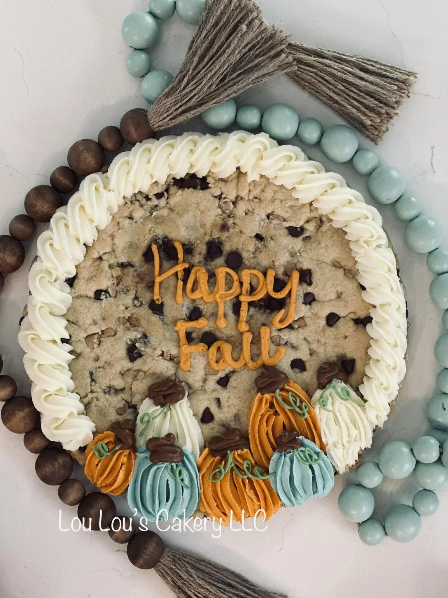 A round fall-themed cookie cake with white icing around the edges, decorated with colorful frosting pumpkins in orange, white, and blue, and topped with the text 'Happy Fall' in orange. The cake is surrounded by a bead garland in light blue and brown