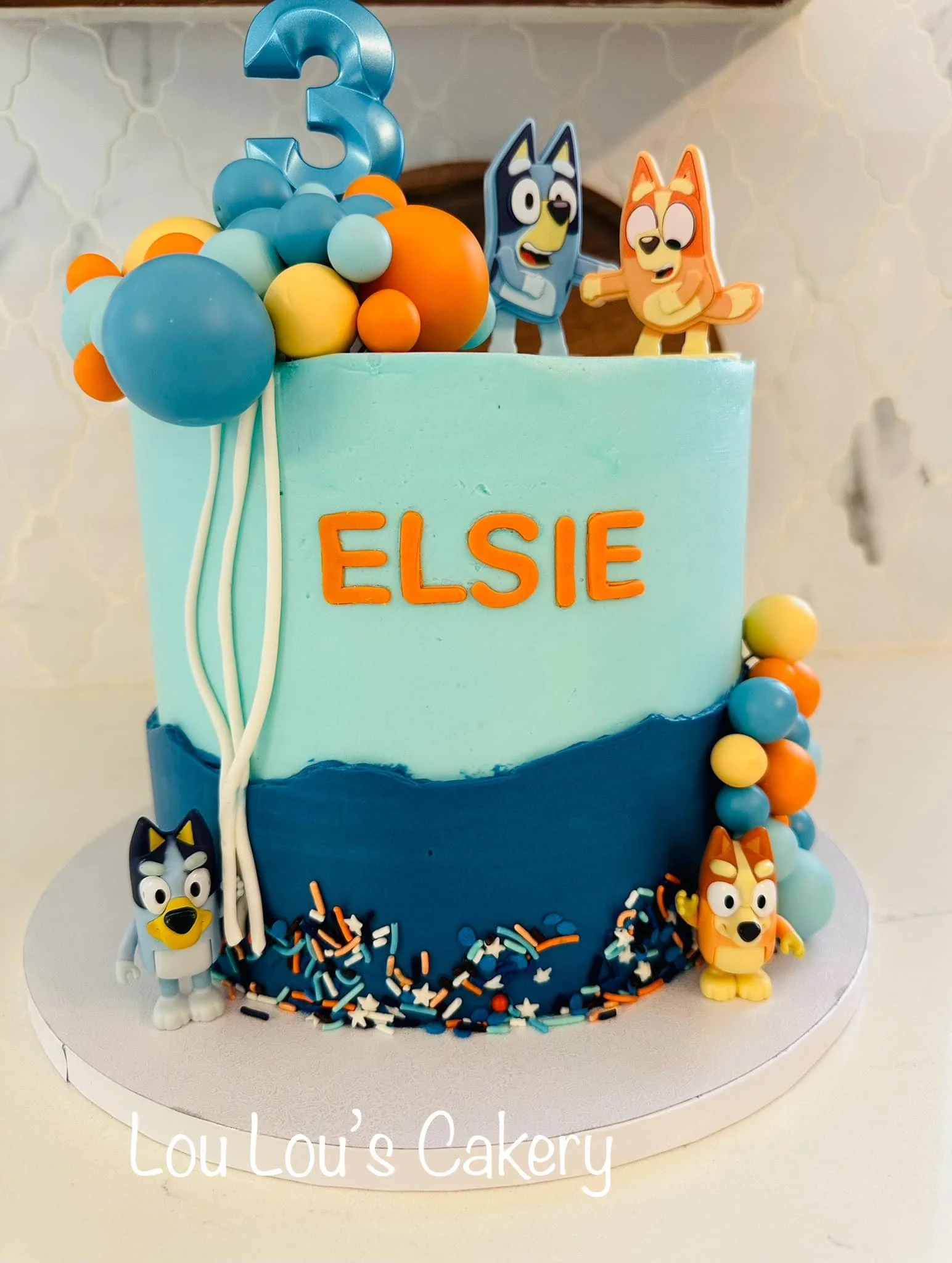 A birthday cake with blue and orange decorations, featuring cartoon dog characters on top, a blue number 3 candle, and the name Elsie in orange letters.