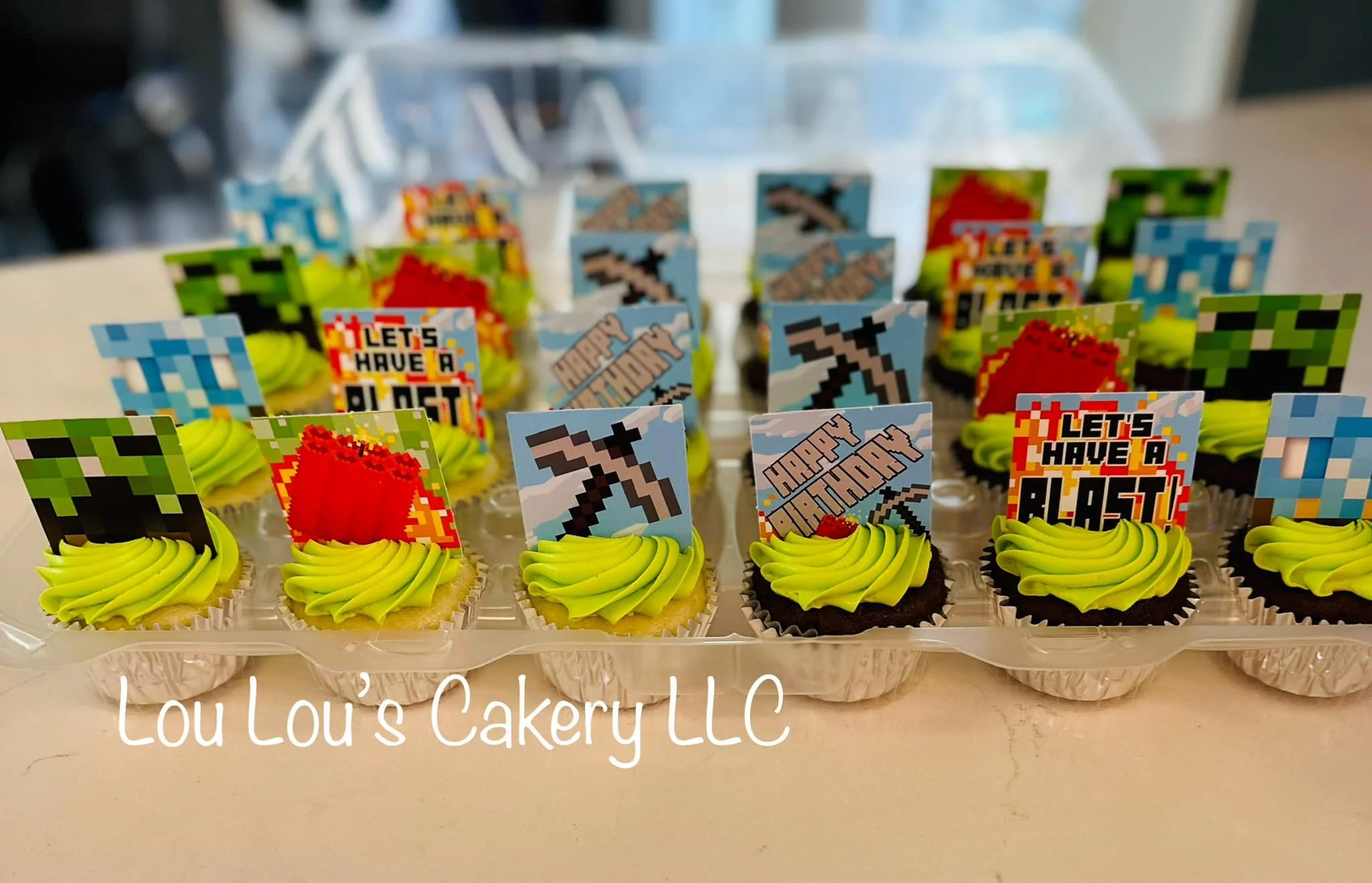 A tray of custom cupcakes decorated with bright green frosting and pixelated video game-themed toppers including swords, pickaxes, TNT blocks, and signs with Minecraft-themed messages. The cupcakes are in silver foil cups, with some chocolate and som