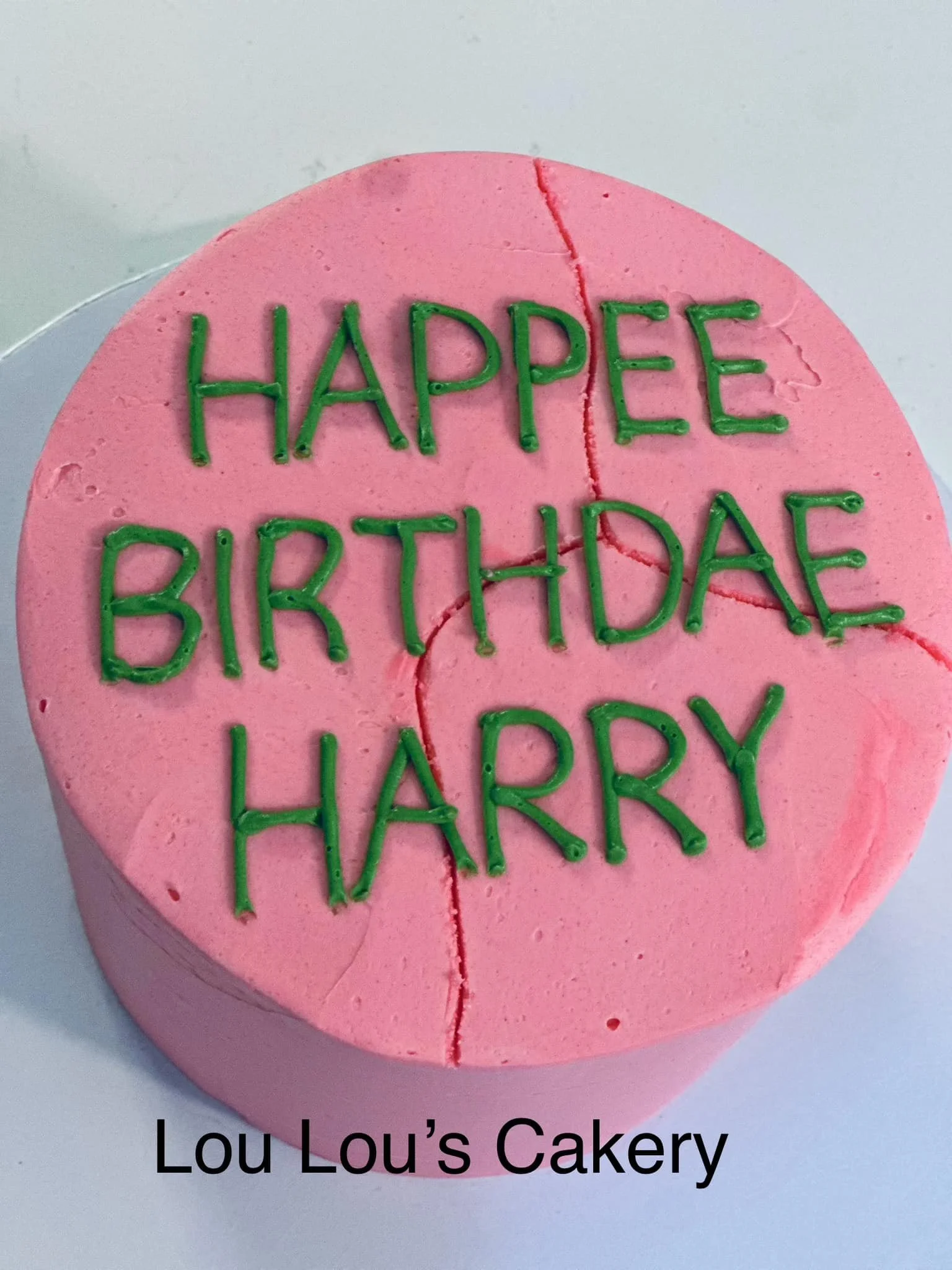 Pink birthday cake with green icing spelling "HAPPEE BIRTHDAE HARRY" and a red crack running from the top to the side, with the text "Lou Lou's Cakery" at the bottom.