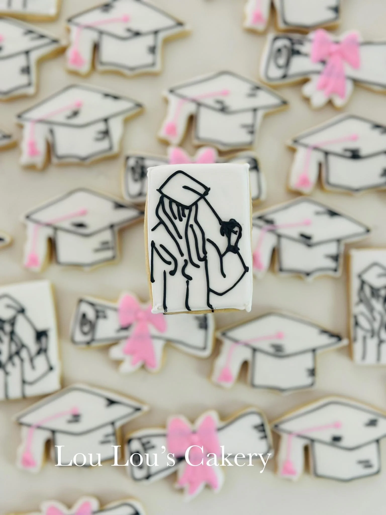 Cookies decorated with graduation cap and gown designs, some with pink bows and ribbons, on a light-colored surface.