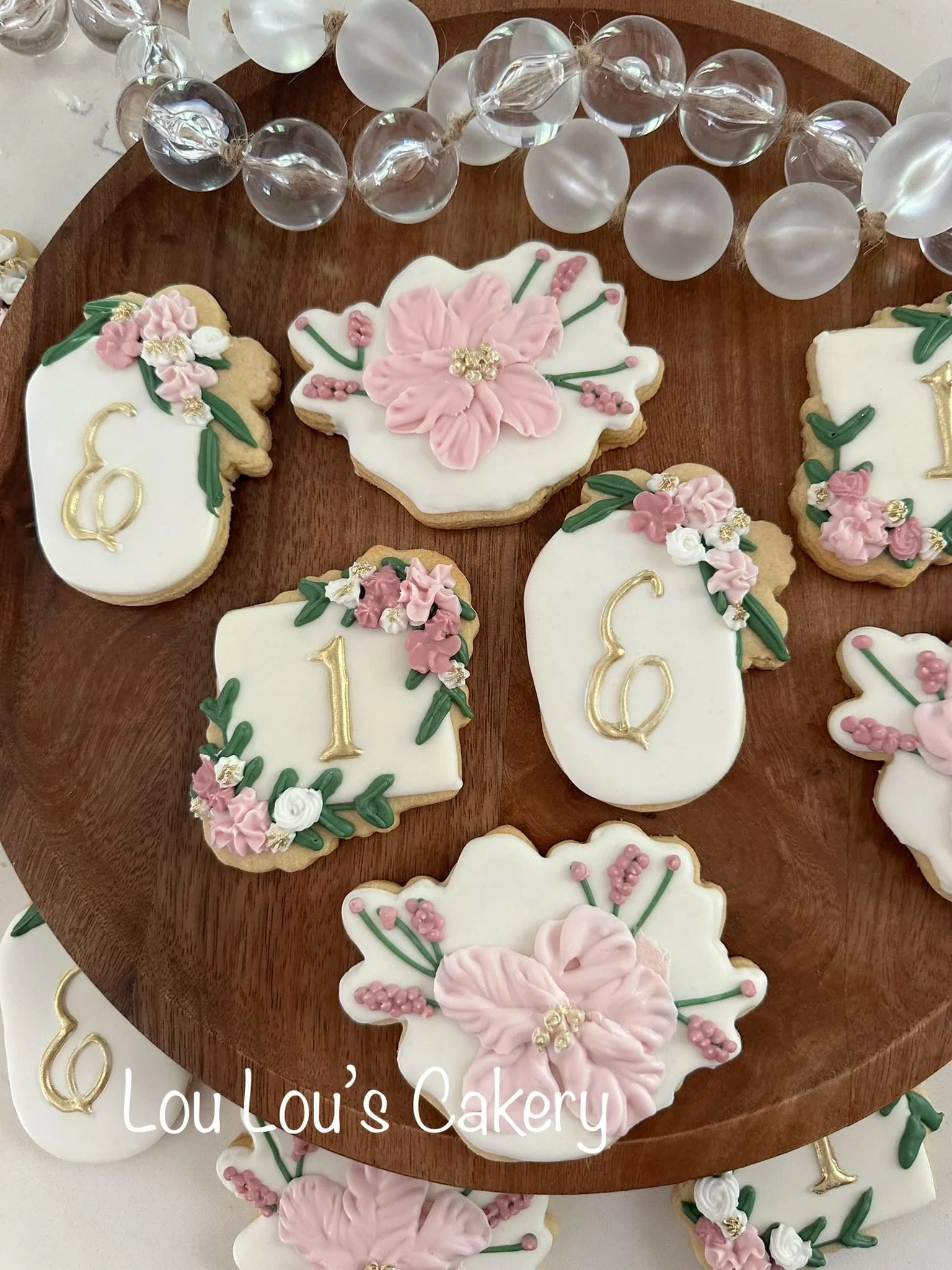 Decorative cookies with white and pink icing, featuring floral designs and gold accents, arranged on a wooden cake stand. Some cookies have the number '1' on them, indicating a celebration, possibly a first birthday or anniversary.