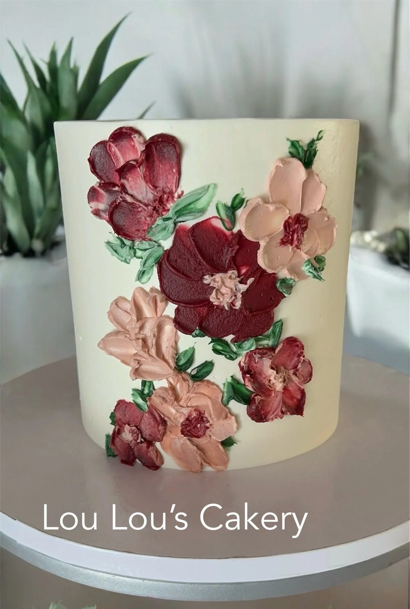 A floral decorated cake with pink and red flowers painted on its surface, placed on a round cake stand with a plant in the background.