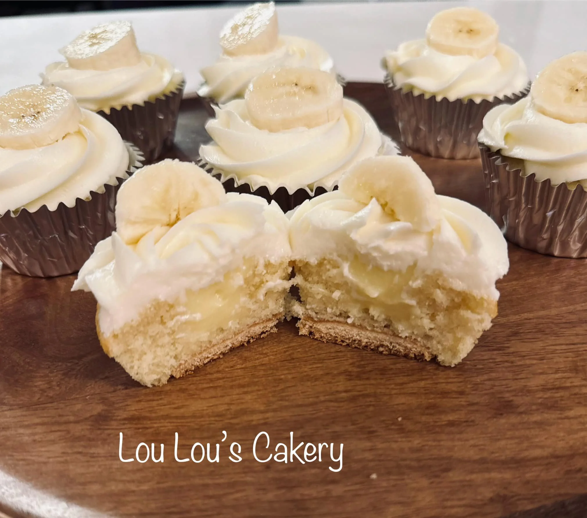 Chocolate cupcakes with vanilla frosting topped with banana slices on a wooden platter, one cut in half showing the filling inside. Text: Lou Lou's Cakery.