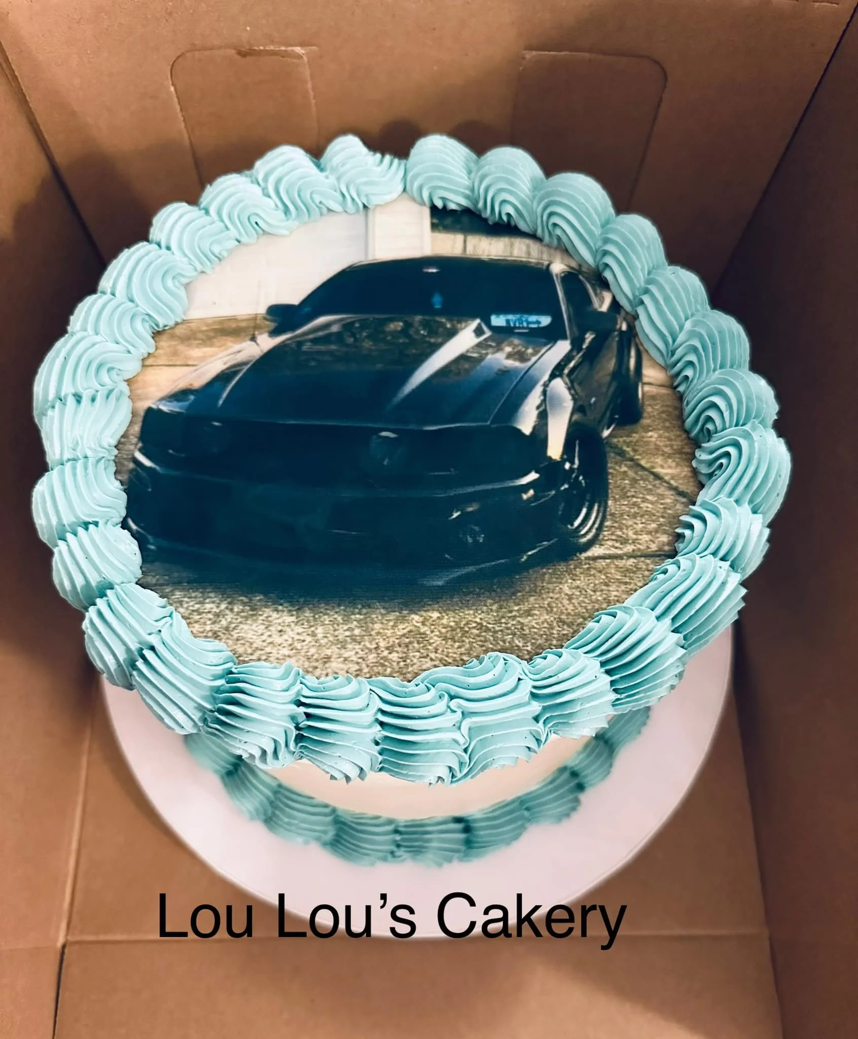 Cake with an edible photo of a black sports car on top, decorated with light blue frosting around the edges, placed inside a cardboard box.