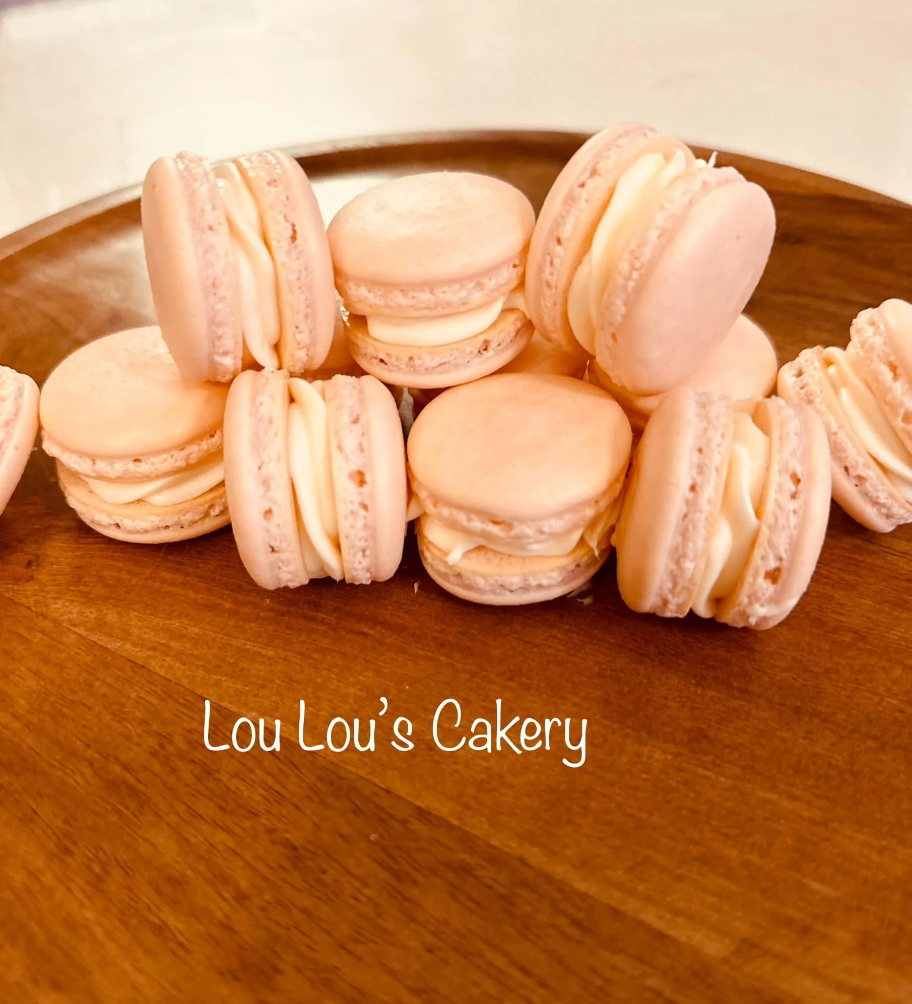 A wooden plate with light pink macarons filled with cream, and the text 'Lou Lou's Cakery' at the bottom.