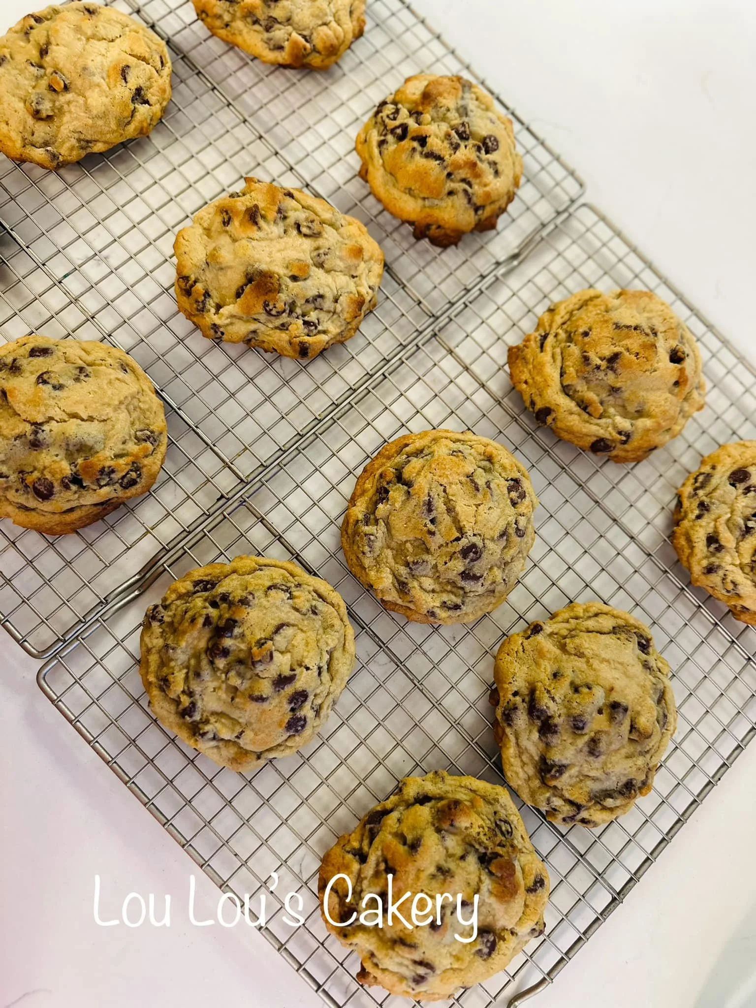 Several freshly baked chocolate chip cookies on cooling racks, with some cookies overlapping. Text overlay says "Lou Lou's Cakery."