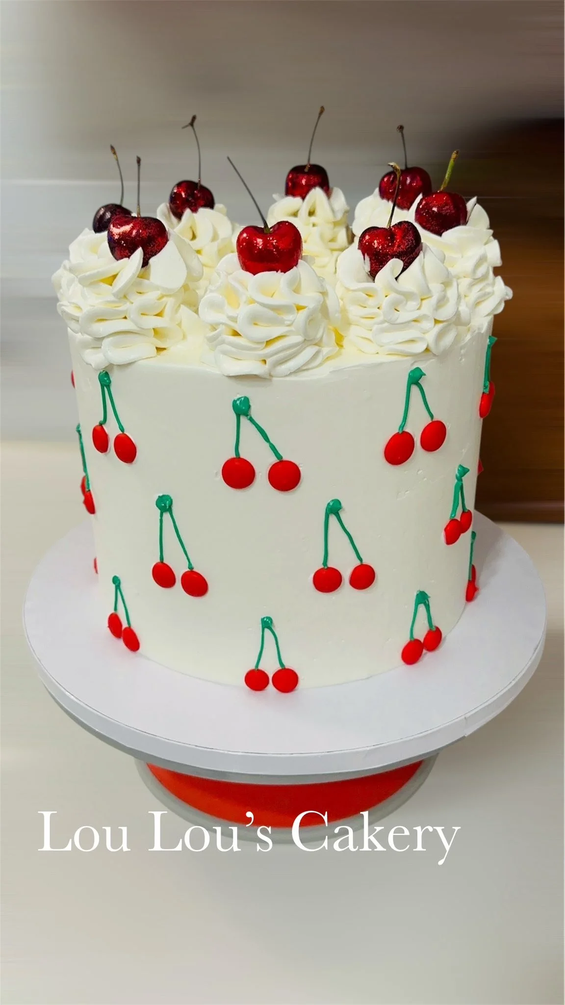 A white frosted cake decorated with whipped cream, cherries, and cherry-themed icing details, displayed on a cake stand.