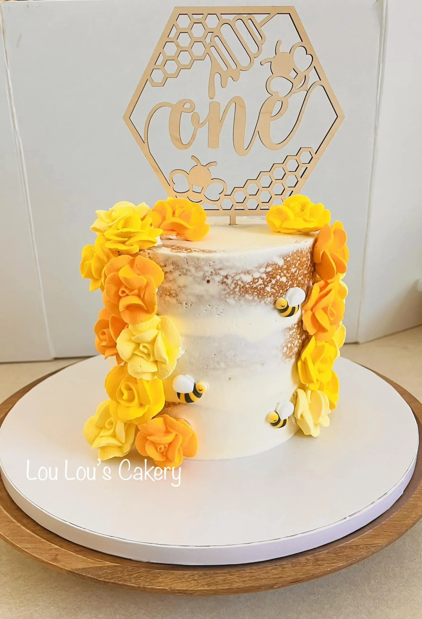 A honey-themed first birthday cake with yellow and orange buttercream flowers, small bee decorations, and a custom cake topper that says 'one' inside a honeycomb design.
