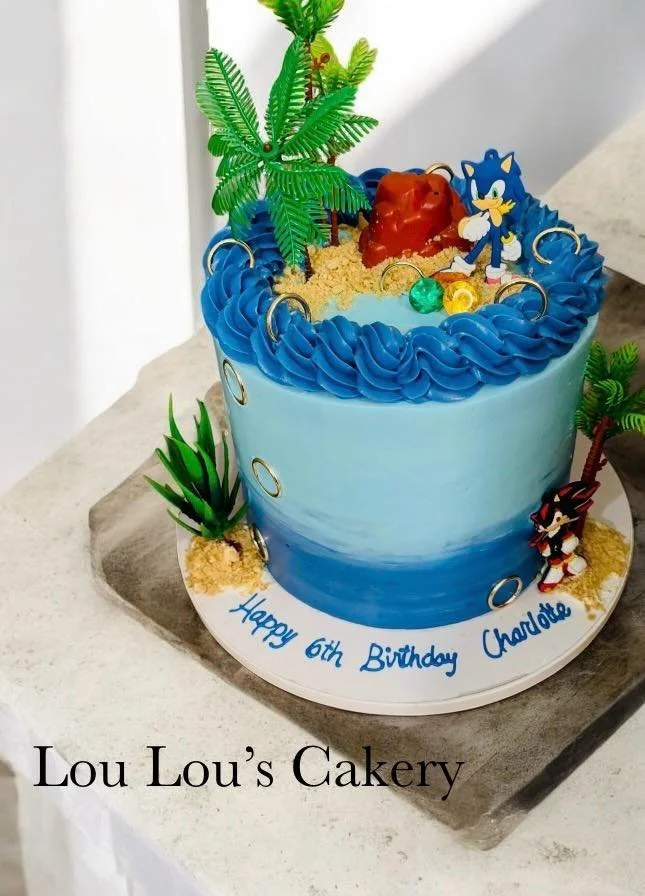 A Sonic the Hedgehog themed birthday cake with blue frosting, palm trees, and Sonic and Tails figurines, celebrating a child's sixth birthday.