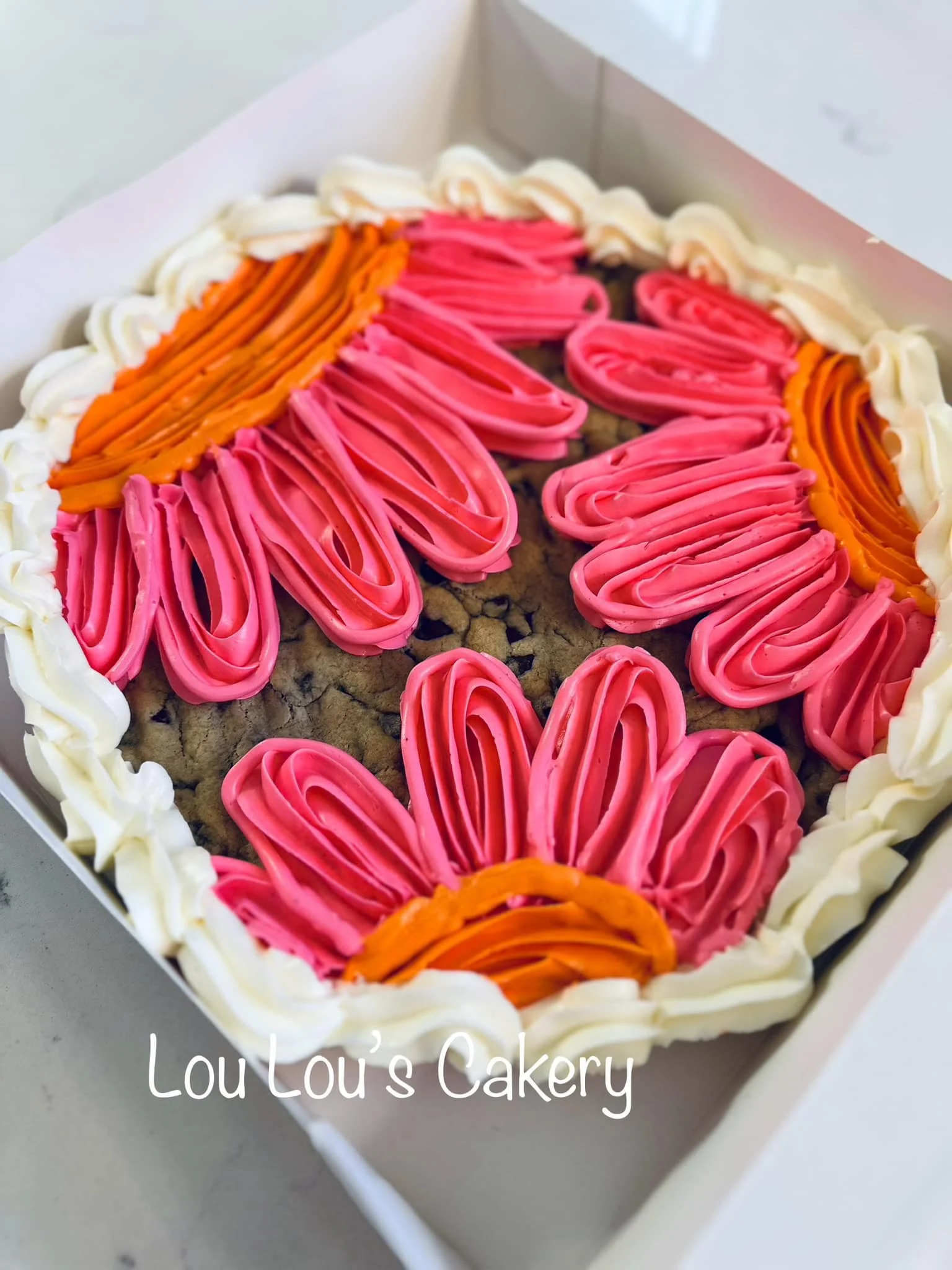 Chocolate chip cookie cake decorated with pink and orange frosting in the shape of flowers, with a white border. The cake has "Lou Lou's Cakery" written on it.