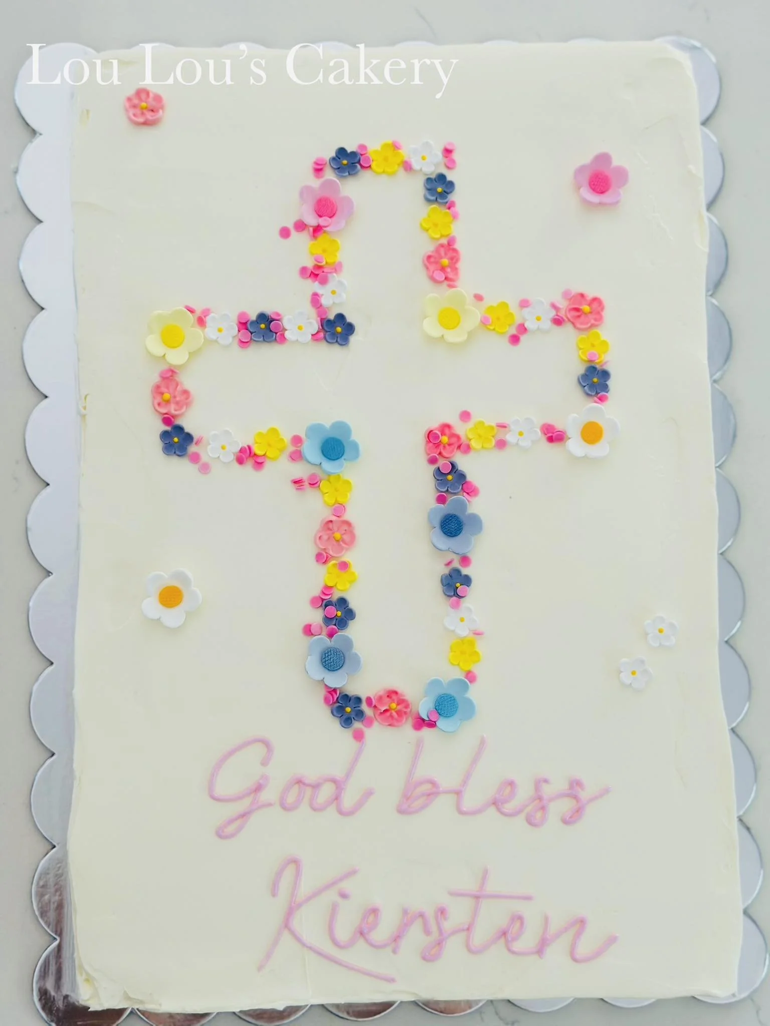 Cake decorated with small colorful flower-shaped sprinkles in the shape of a cross, with the message "God bless Kiersten" written in pink icing.
