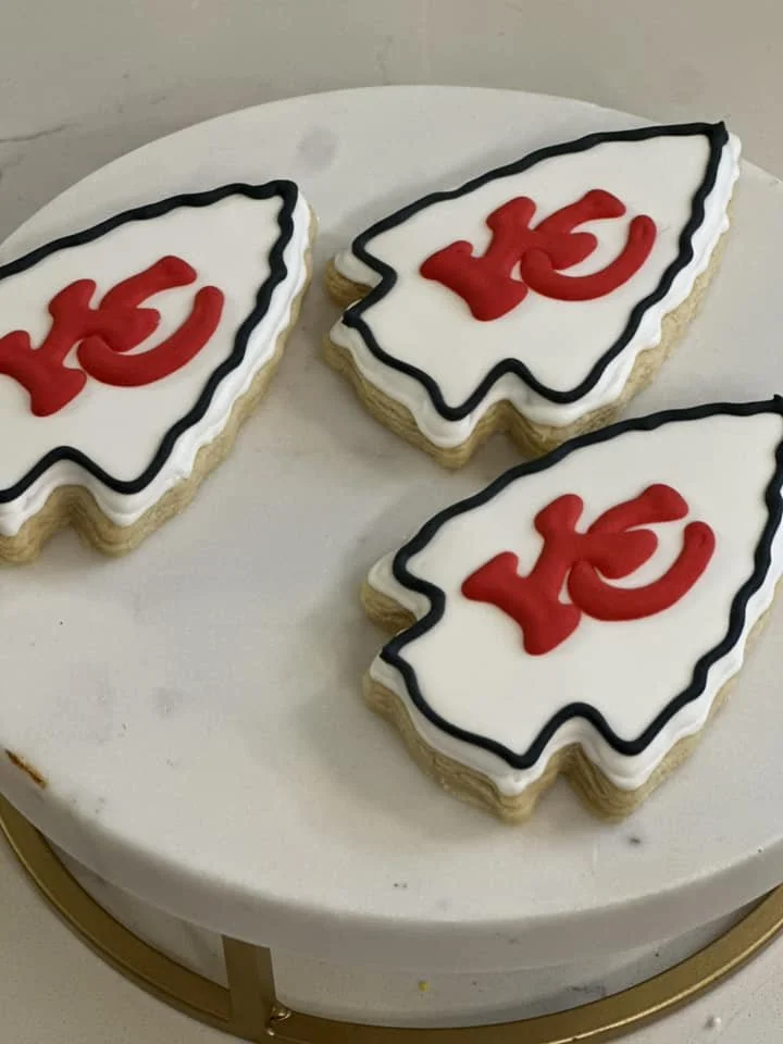 Three cookies decorated with white icing, red 'KC' logos, and black outline, on a white fondant-covered cake.