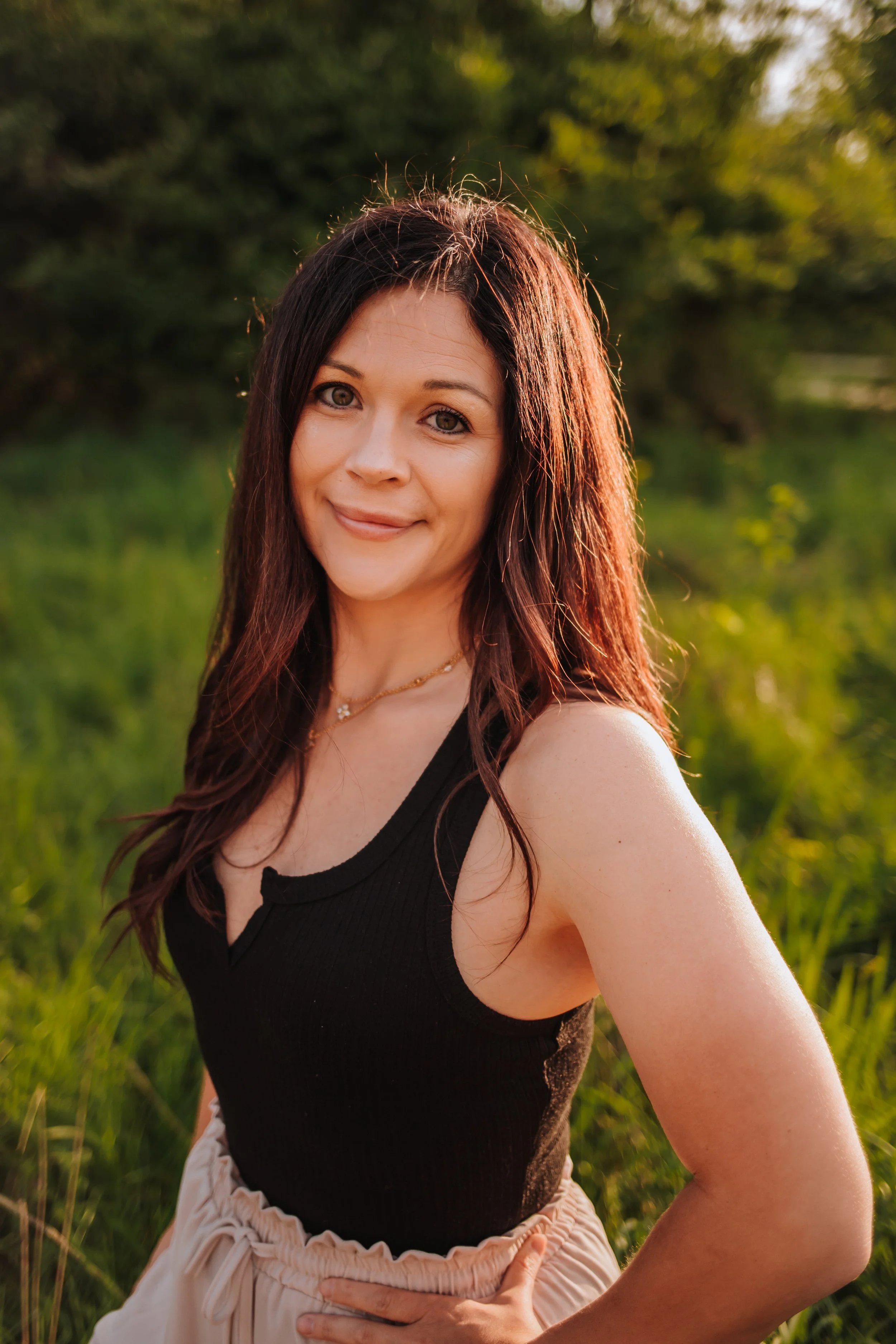 A woman with long reddish-brown hair smiling outdoors in a green field during sunset.