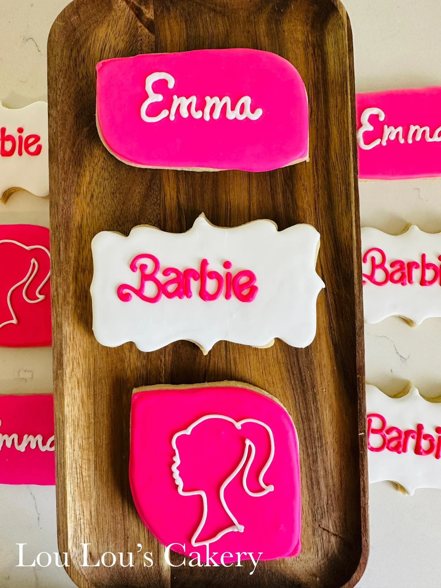 Assorted decorated cookies on a wooden tray, featuring pink and white icing with the words 'Emma' and 'Barbie' and a silhouette of a girl's profile with a ponytail.