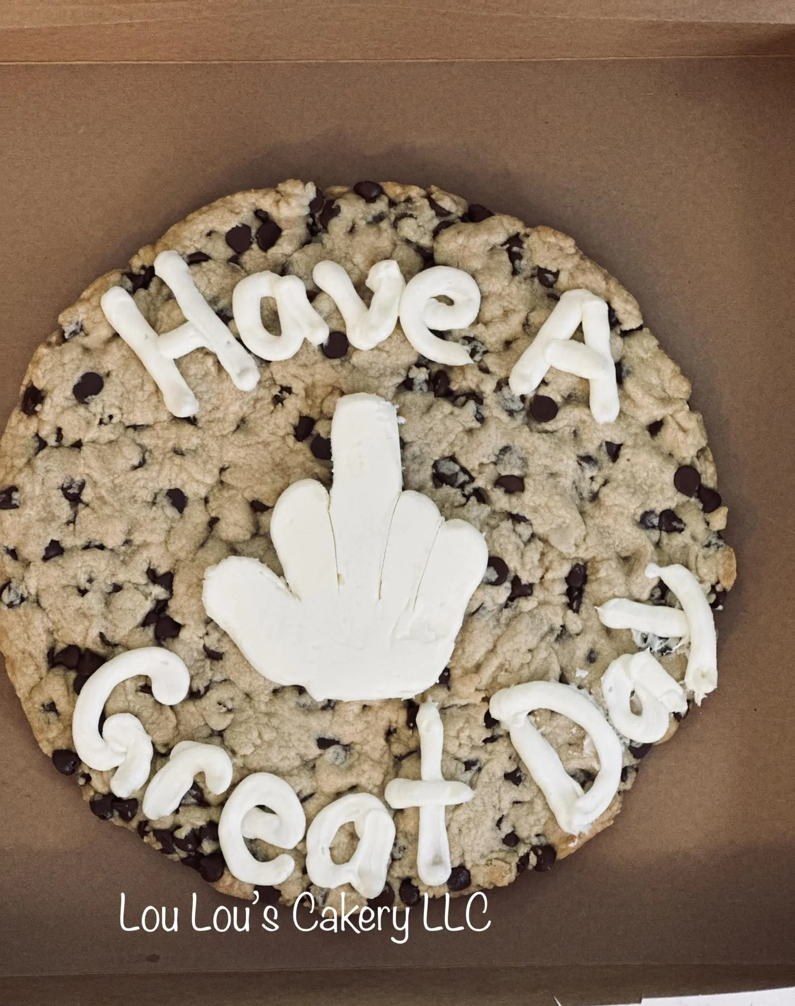 Chocolate chip cookie cake with white icing letters spelling 'Have A Great Day' and a hand making the middle finger gesture, with 'Lou Lou's Cakery LLC' written at the bottom.