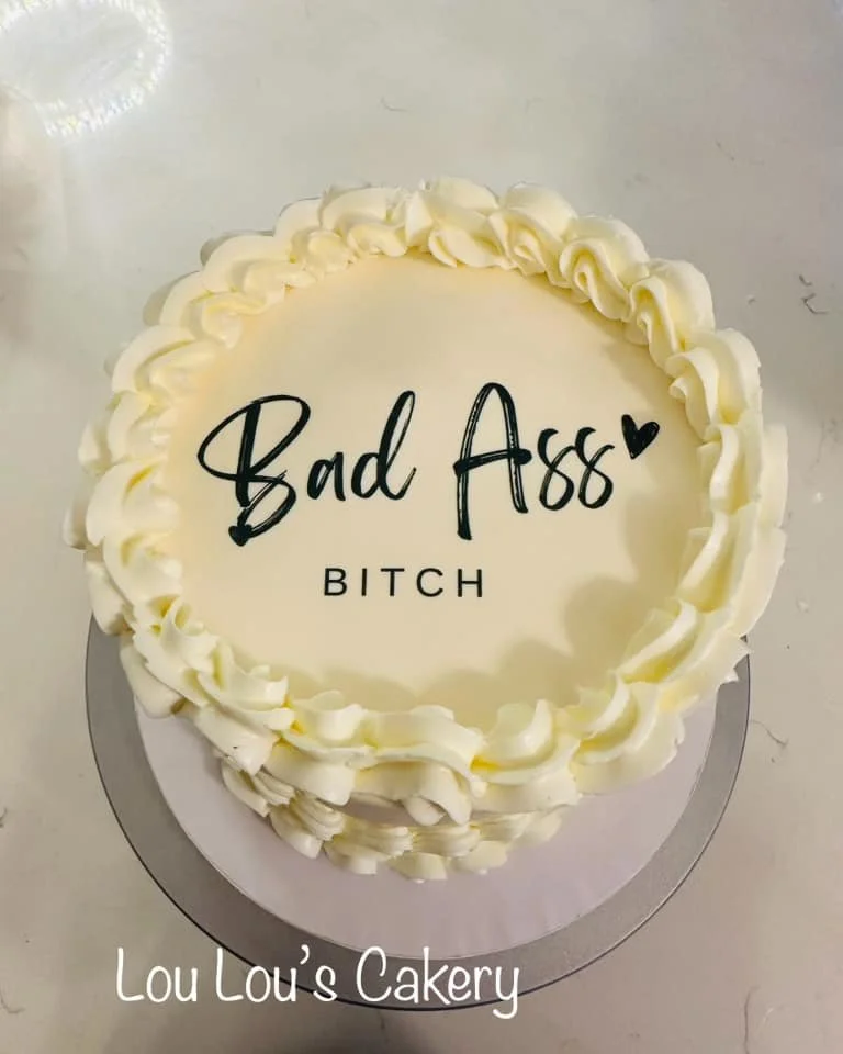 A round white cake with white frosting decorated with a swirl border, displaying the message 'Bad Ass Bitch' in black lettering with a small black heart.