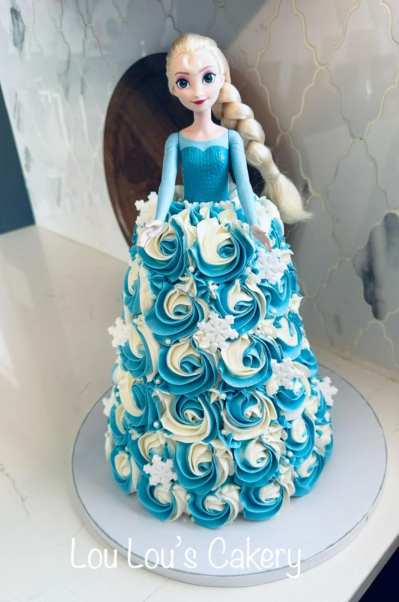 A cake designed to look like Elsa from Frozen, with a doll on top wearing a blue dress and a skirt made of blue and white rosette frosting, decorated with snowflakes.
