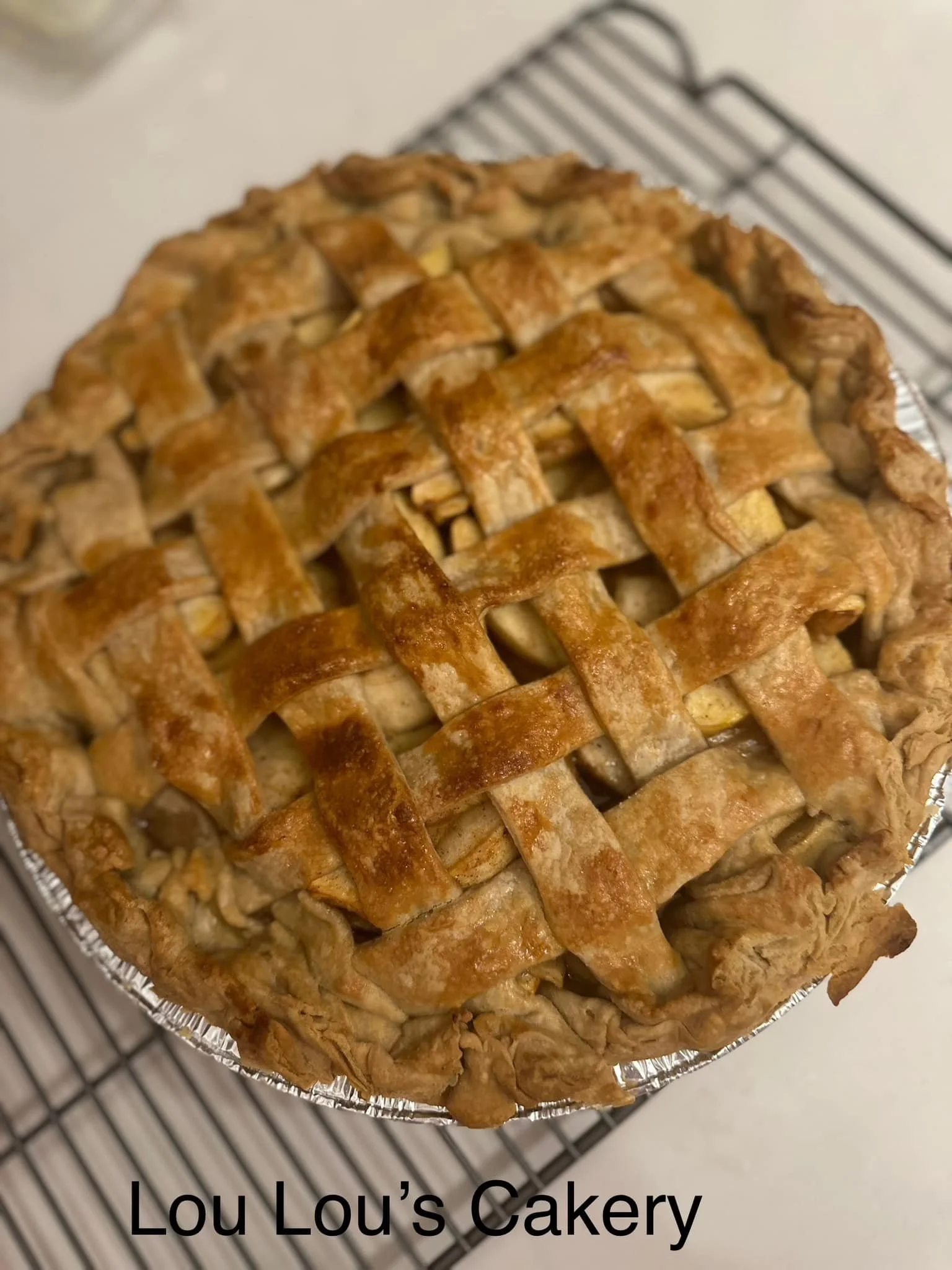 A homemade apple pie with a lattice crust cooling on a wire rack. The pie is in a disposable aluminum pie pan, and the crust has a golden-brown color.