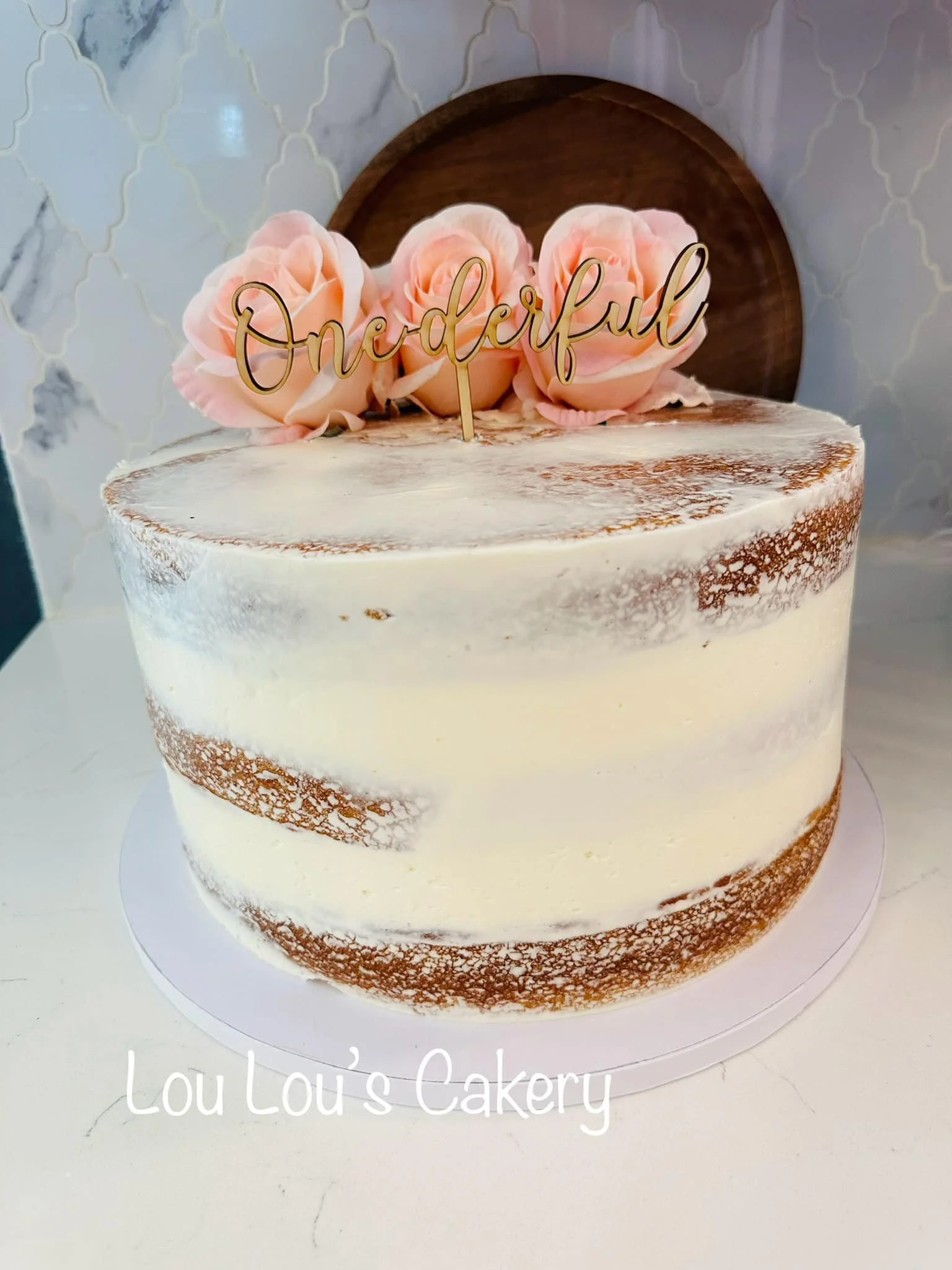 A semi-naked cake with white frosting, decorated with pink roses and a gold "One Diful" topper, on a white surface. A wooden cake topper and pink roses are on top of the cake, with a marble-patterned wall in the background.