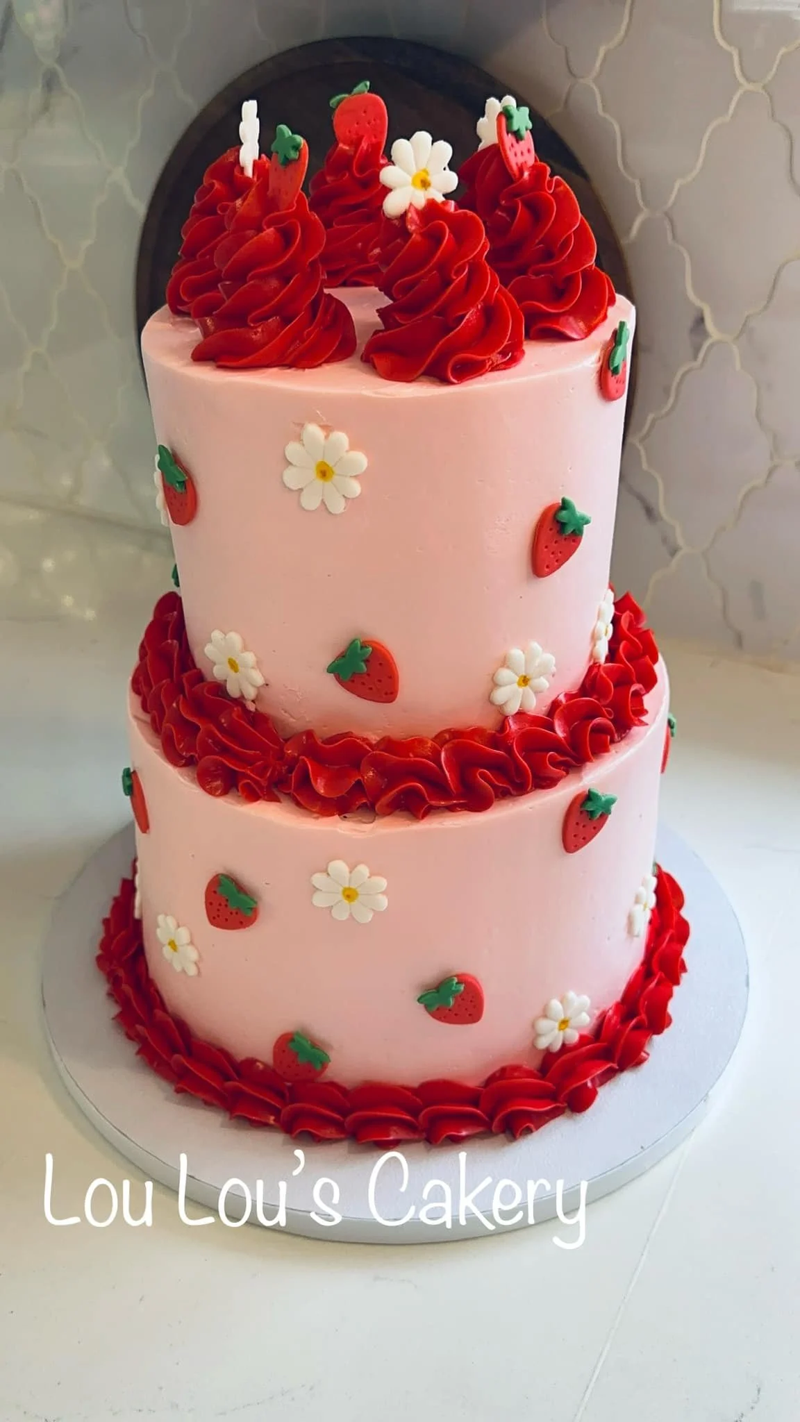 A three-tier pink cake decorated with strawberries, white flowers, and red icing, with red rosettes on top, placed on a round white cake board.