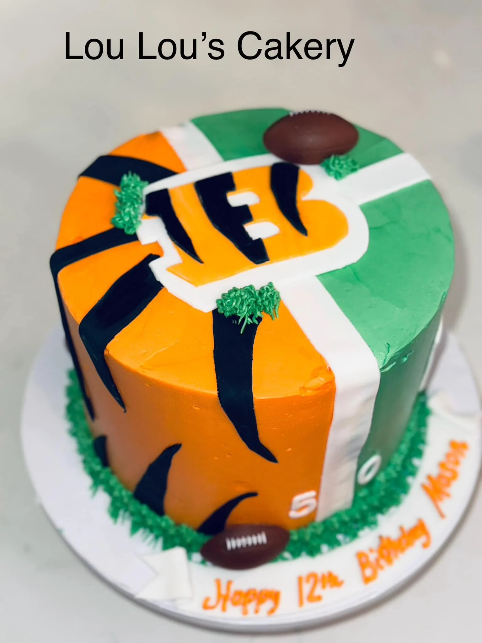 A colorful, football-themed cake celebrating a 12th birthday. The cake has orange, green, and white sections with black stripes, a football topper, and the number 12 in orange. The cake's message reads, "Happy 12th Birthday Mayson," in orange icing o