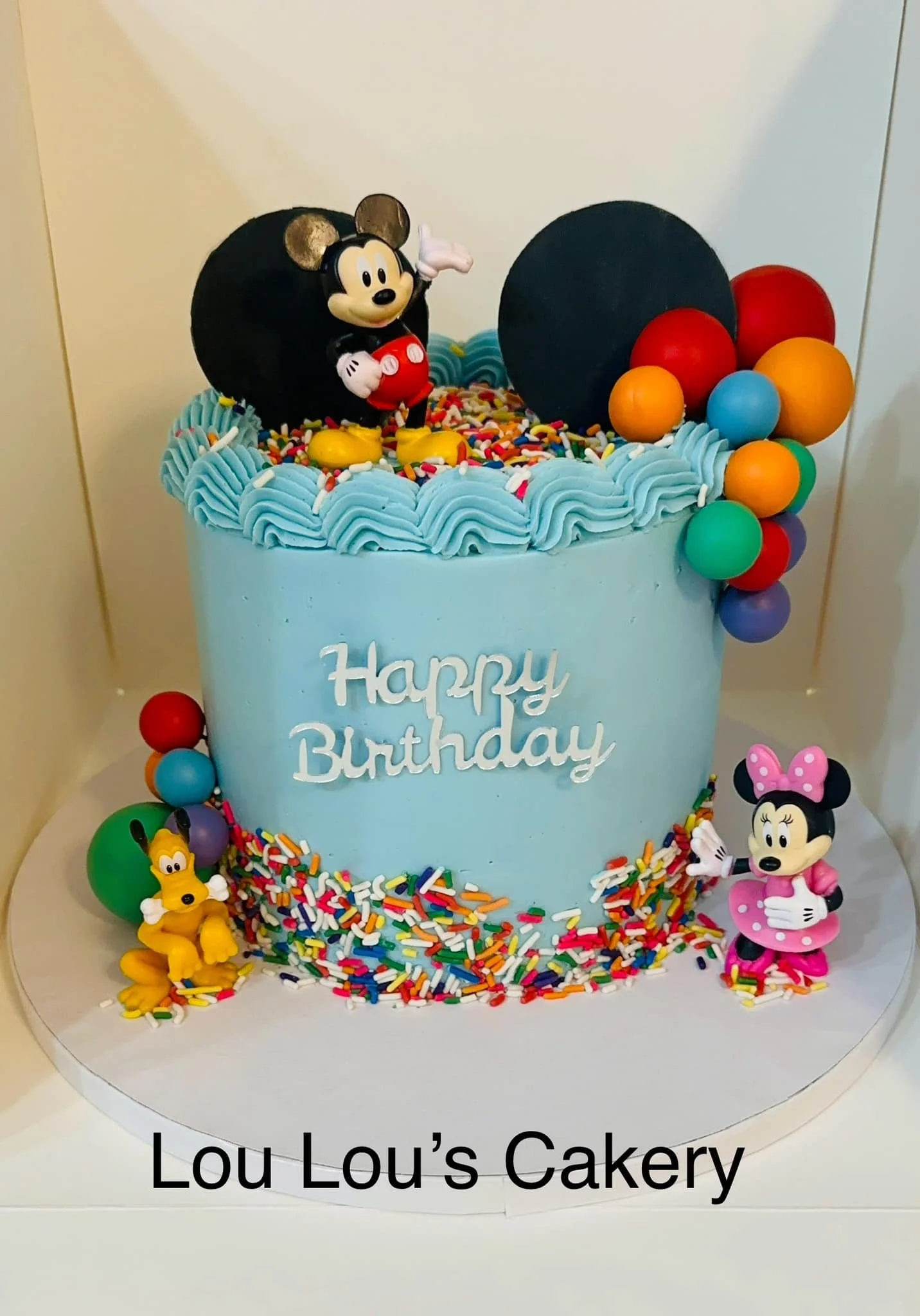 A birthday cake with blue frosting, decorated with colorful sprinkles, featuring a Mickey Mouse figurine on top and Minnie Mouse and Pluto figurines at the base. The cake has the message 'Happy Birthday' written on it and is surrounded by multicolore