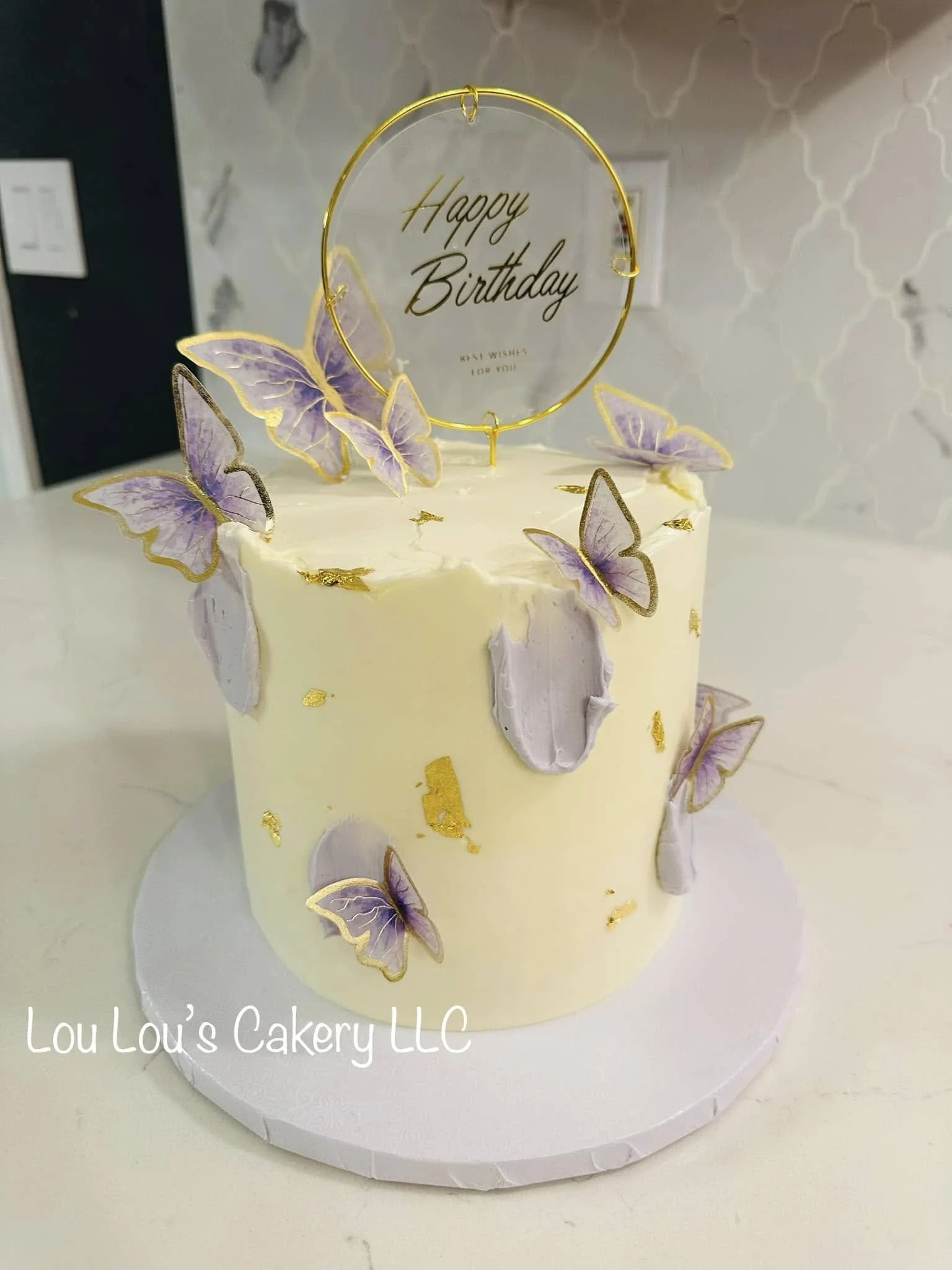 A white birthday cake decorated with purple and gold butterfly-shaped toppers, with a round, gold wire 'Happy Birthday' topper on top. The cake sits on a white cake board with the words 'Lou Lou's Cakery LLC' written on it.