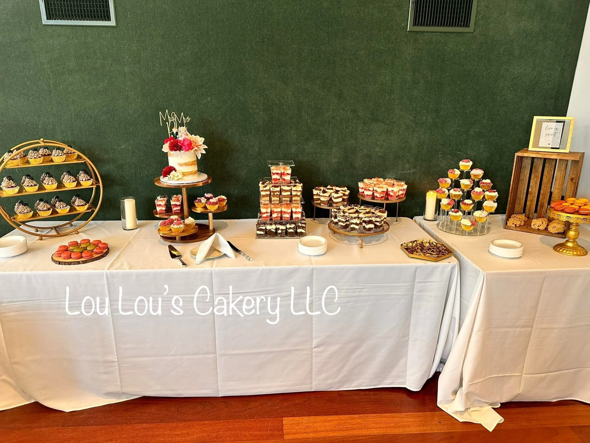 Dessert table with cupcakes, layered cakes, parfaits, and cookies at Lou Lou's Cakery LLC event, decorated with candles and a dark green wall background.