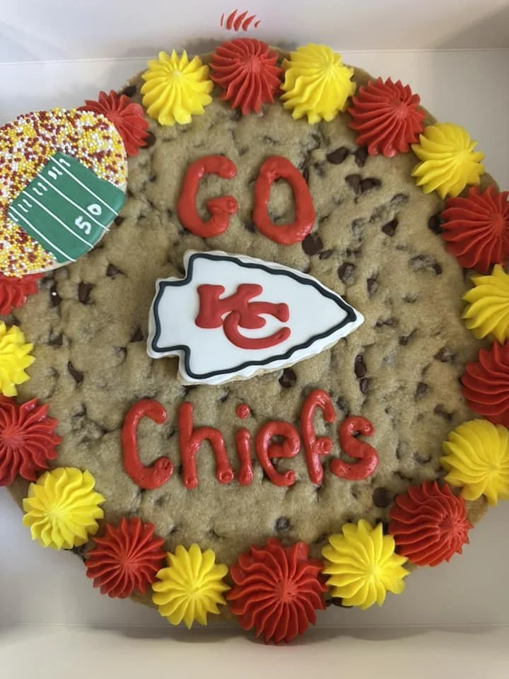 A large cookies decorated with red and yellow icing flowers, a white football-shaped cookie with a black outline, and the words 'GO' and 'Chiefs' spelled out in red icing. The cookie has chocolate chips inside.