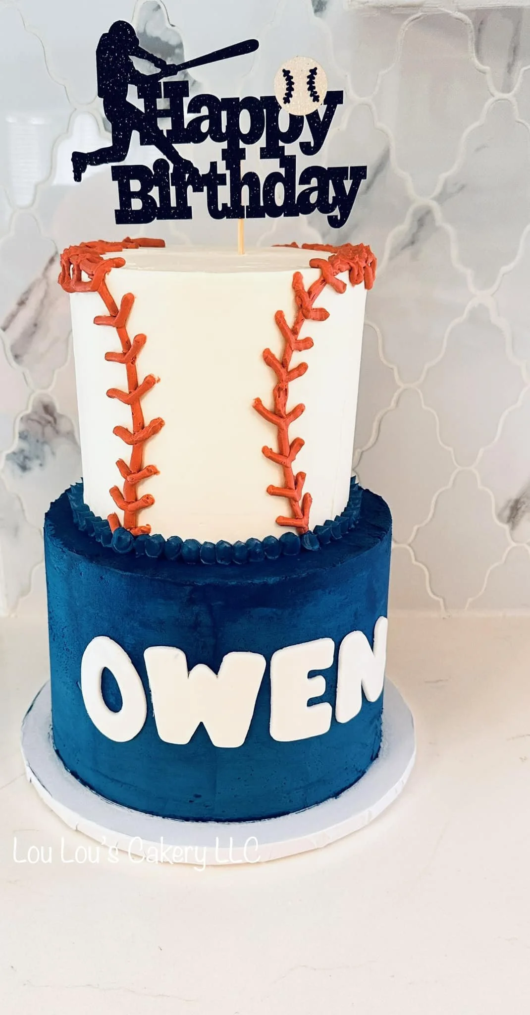 Two-tiered birthday cake with baseball theme, featuring a navy blue bottom layer with white letters spelling OWIN, and a white top layer decorated with baseball stitching, topped with a baseball and a silhouette of a baseball player swinging a bat sa