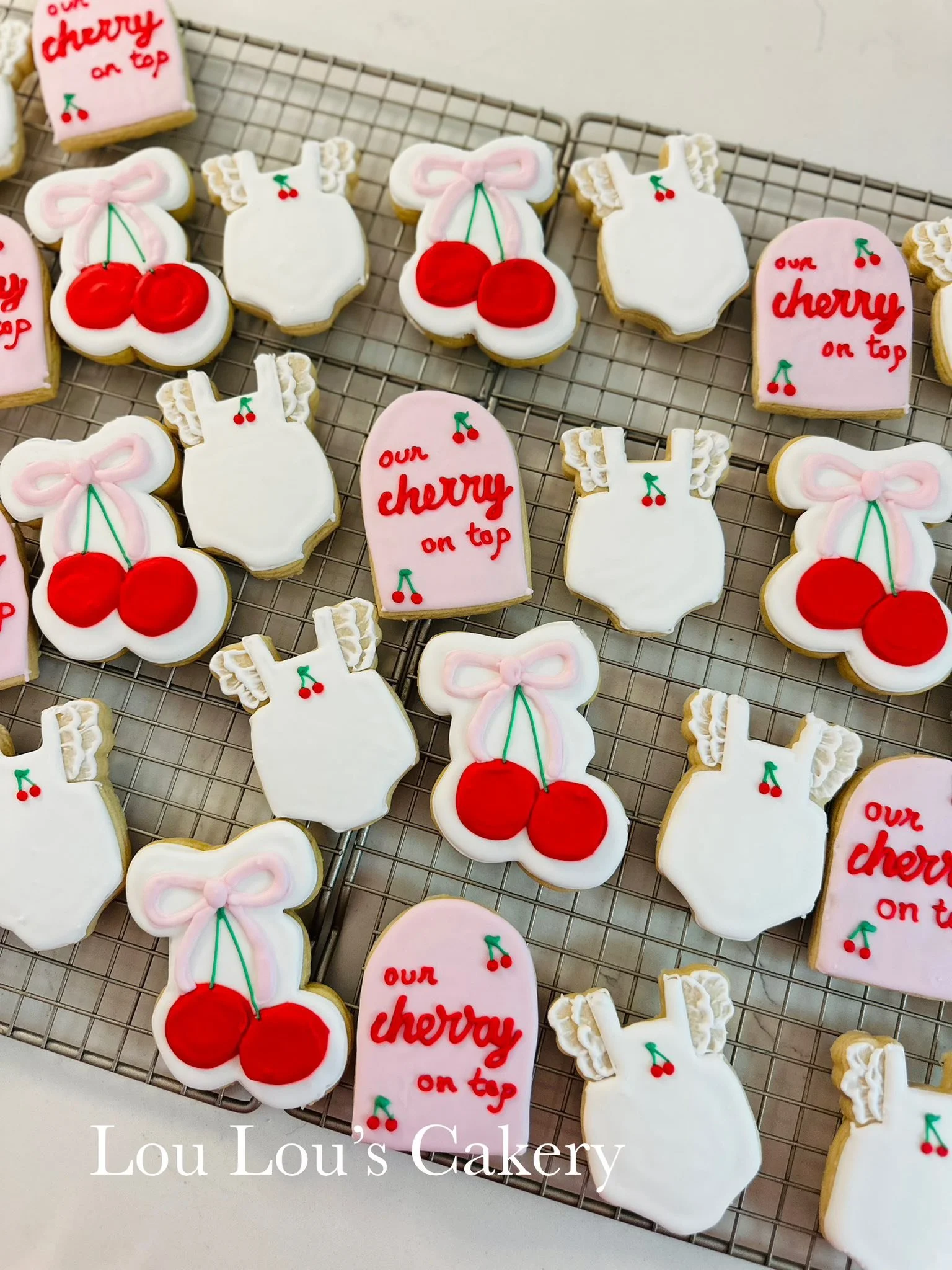 Decorated cookies in cherry and dress shapes with red, white, and pink icing, on a cooling rack, with text indicating 'Lou Lou's Cakery'.