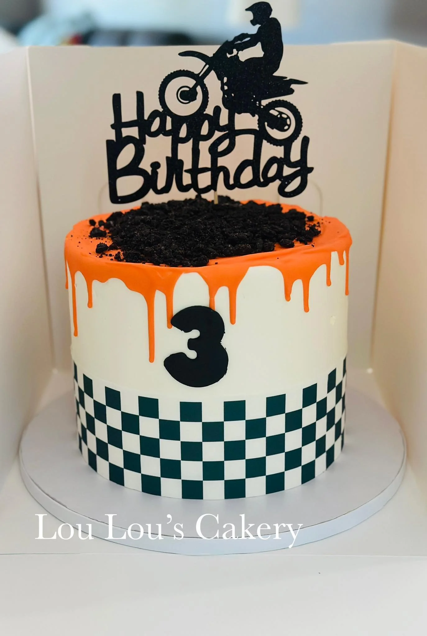 A birthday cake decorated with racing theme elements. The cake has white icing with a black and white checkered border, orange drip icing on top, crushed cookies resembling dirt, and a "Happy Birthday" topper with a silhouette of a person riding a di