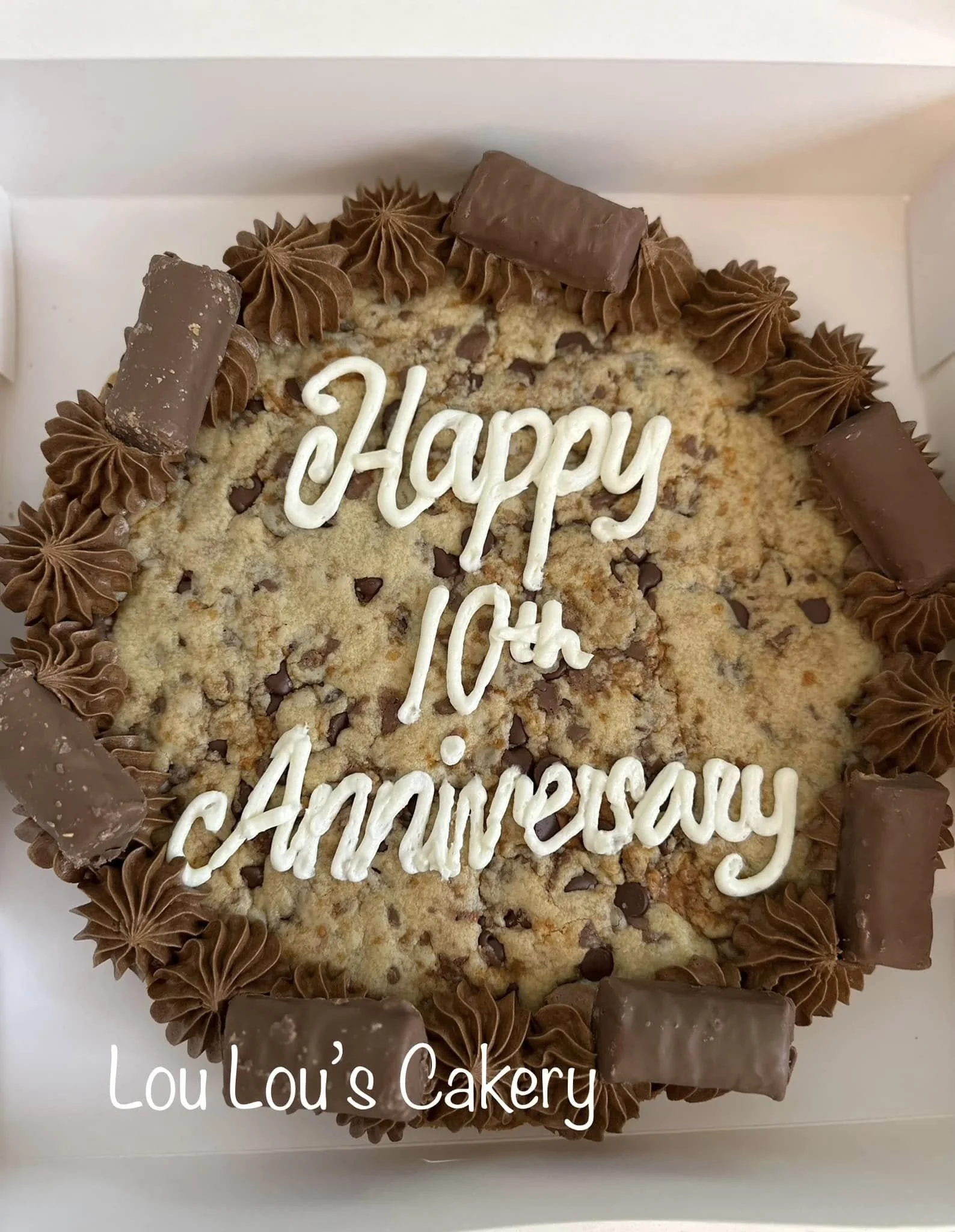 Chocolate chip cookie cake with chocolate candies and piped chocolate frosting, decorated for a 10th anniversary, with white script reading 'Happy 10th Anniversary' and the bakery's name, Lou Lou's Cakery.