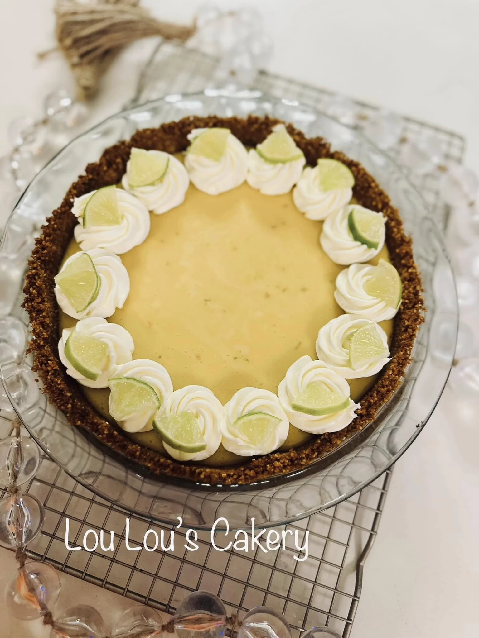 Lemon cheesecake with a graham cracker crust, topped with whipped cream rosettes and lime wedges, displayed on a glass plate.