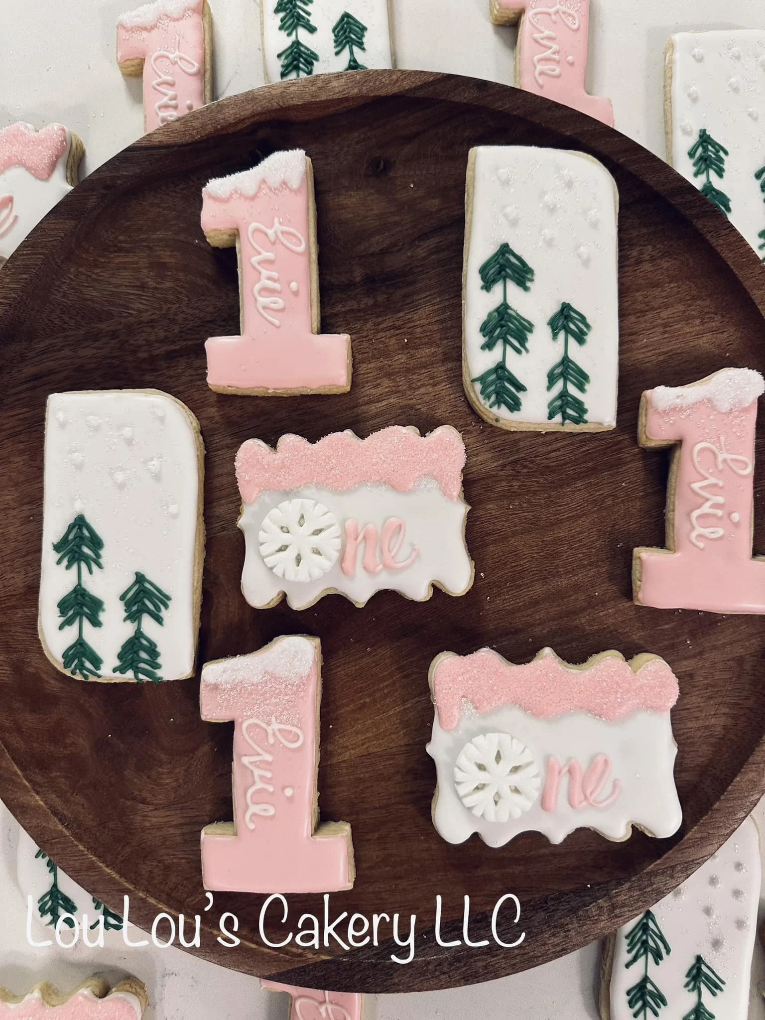 Decorated cookies with pink and white icing, featuring the word "one" and Christmas tree designs, arranged on a wooden tray.
