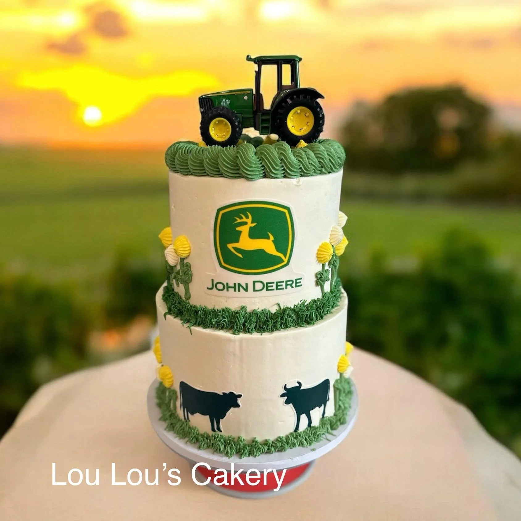 A three-tiered white cake with green and yellow decorations, featuring a John Deere logo and tractor topper, and black silhouette cows on the bottom tier, displayed on a white surface with a countryside sunset background.
