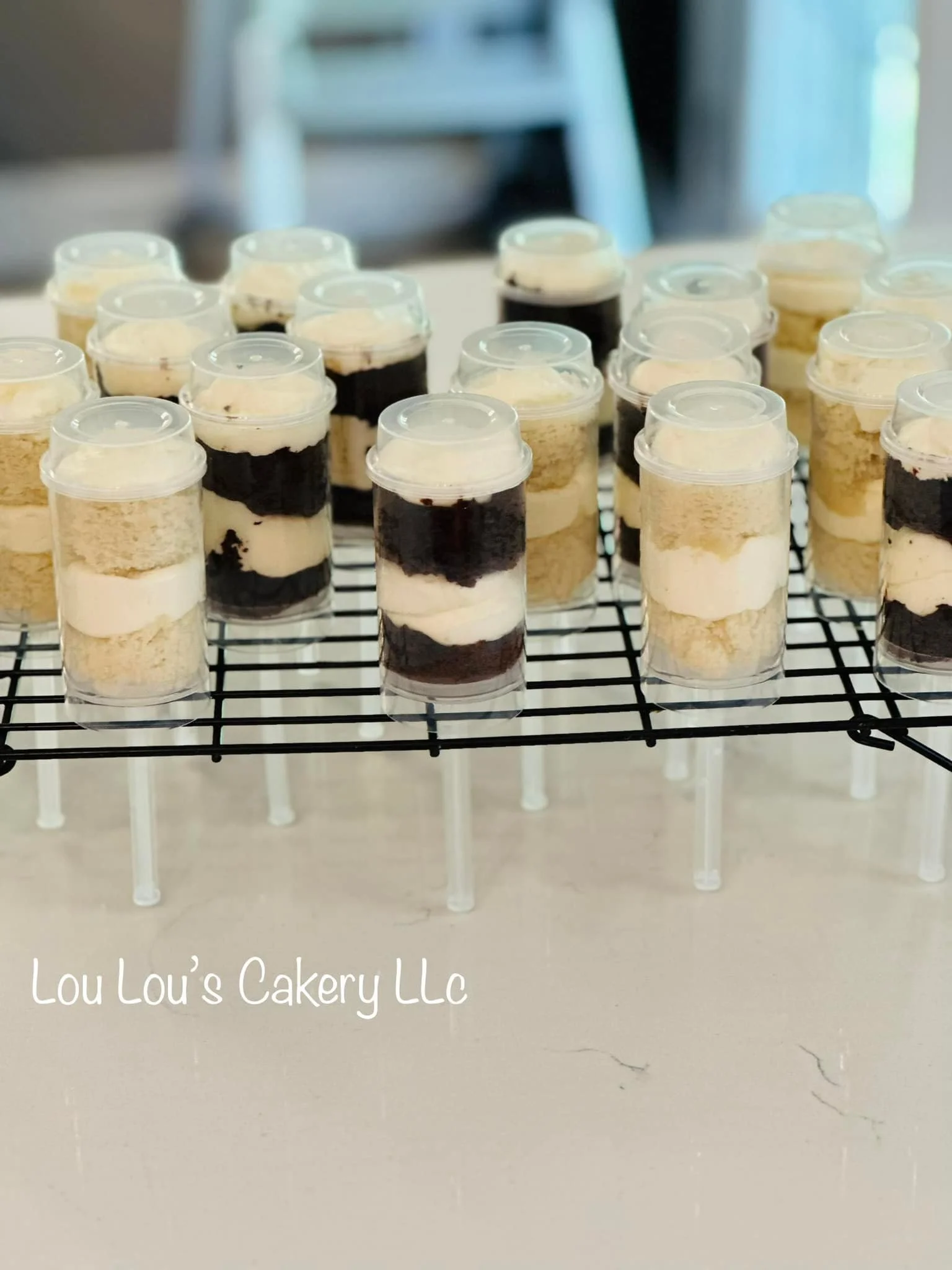 Miniature layered cheesecakes in small clear cups with plastic lids, arranged on a black cooling rack, branded with 'Lou Lou's Cakery LLC' at the bottom left corner.