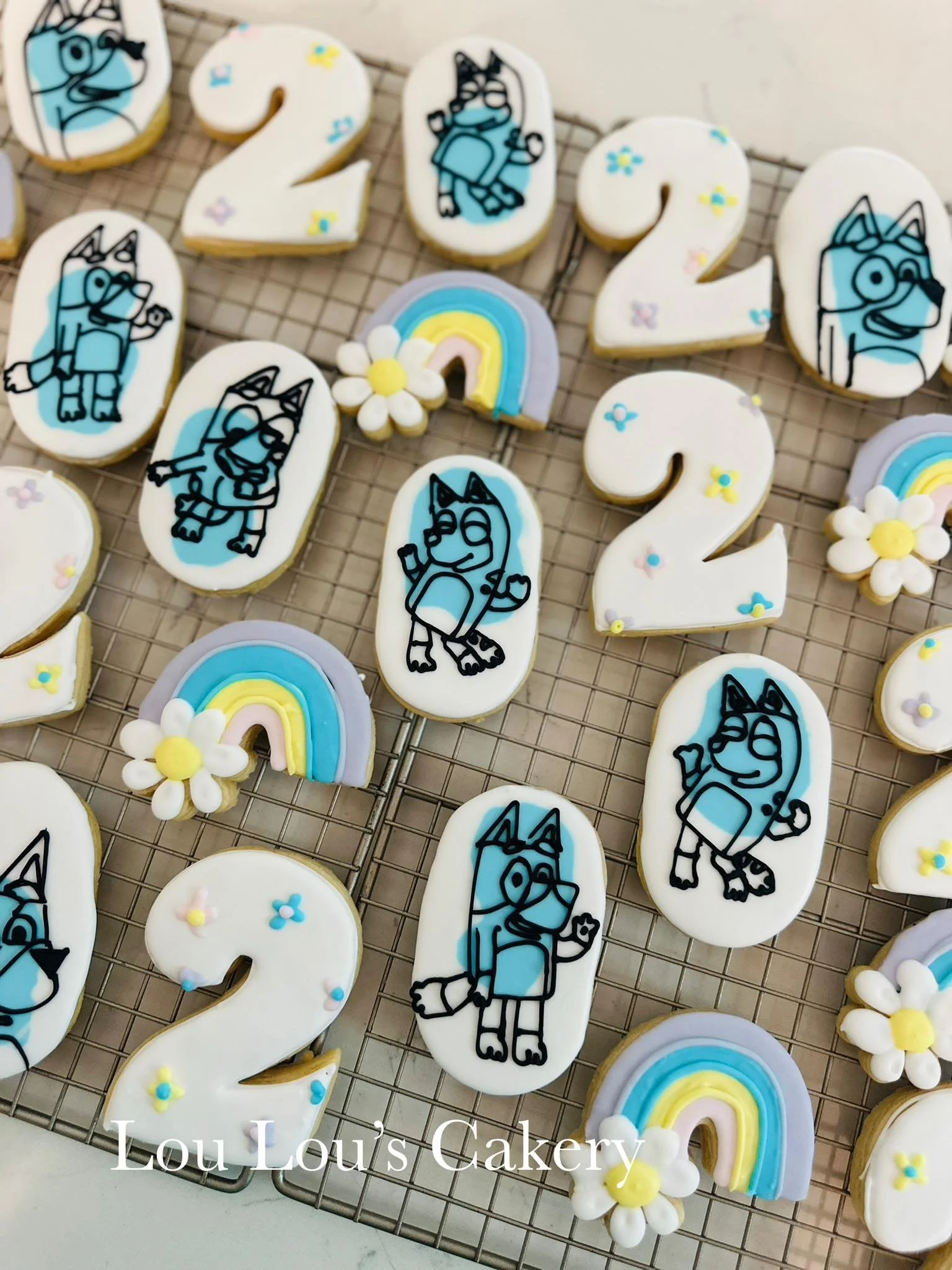 Decorated cookies on a cooling rack featuring a cartoon blue dog, white number cookies with colorful sprinkles, rainbow and flower designs, and the text 'Lou Lou's Cakery'.