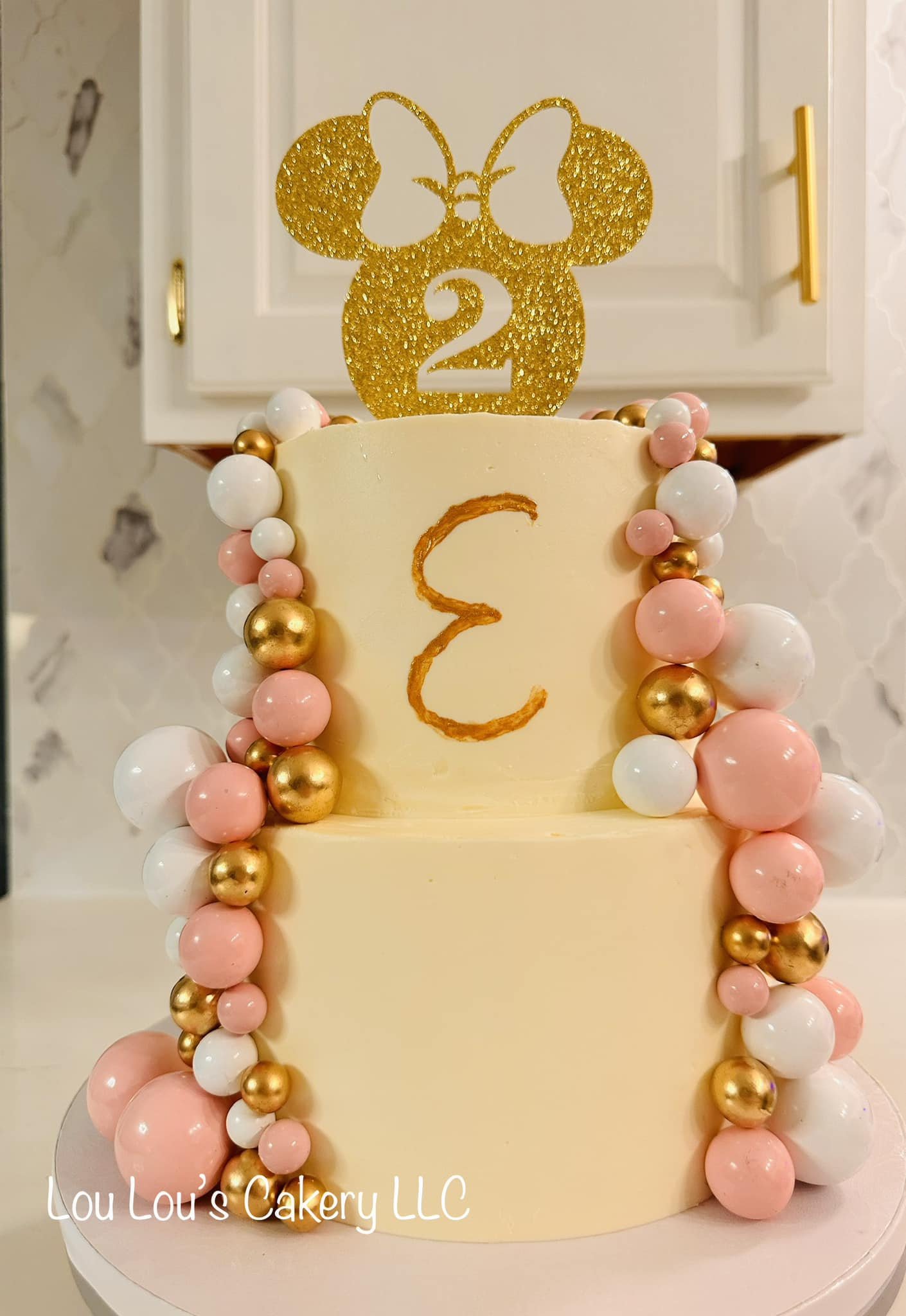Two-tiered cake with yellow fondant, decorated with pink, white, and gold ball ornaments on the sides. The top of the cake has a glittery gold Minnie Mouse figure with a bow and the number '2' in the center. The front of the cake features a letter 'E