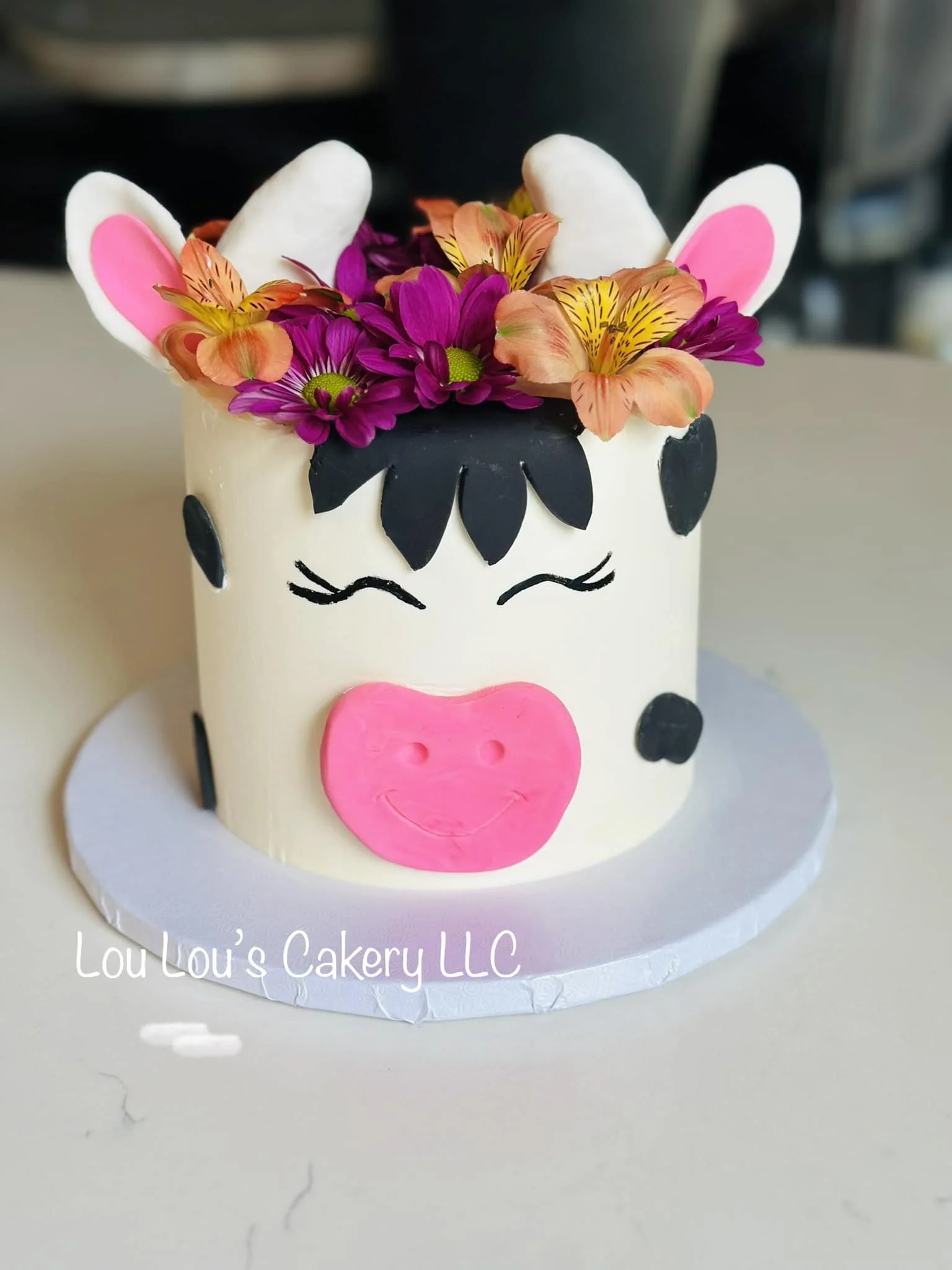 Cake decorated as a cartoon cow with pink and black facial features, topped with colorful flowers and white candy ears with pink centers, displayed on a white round cake board