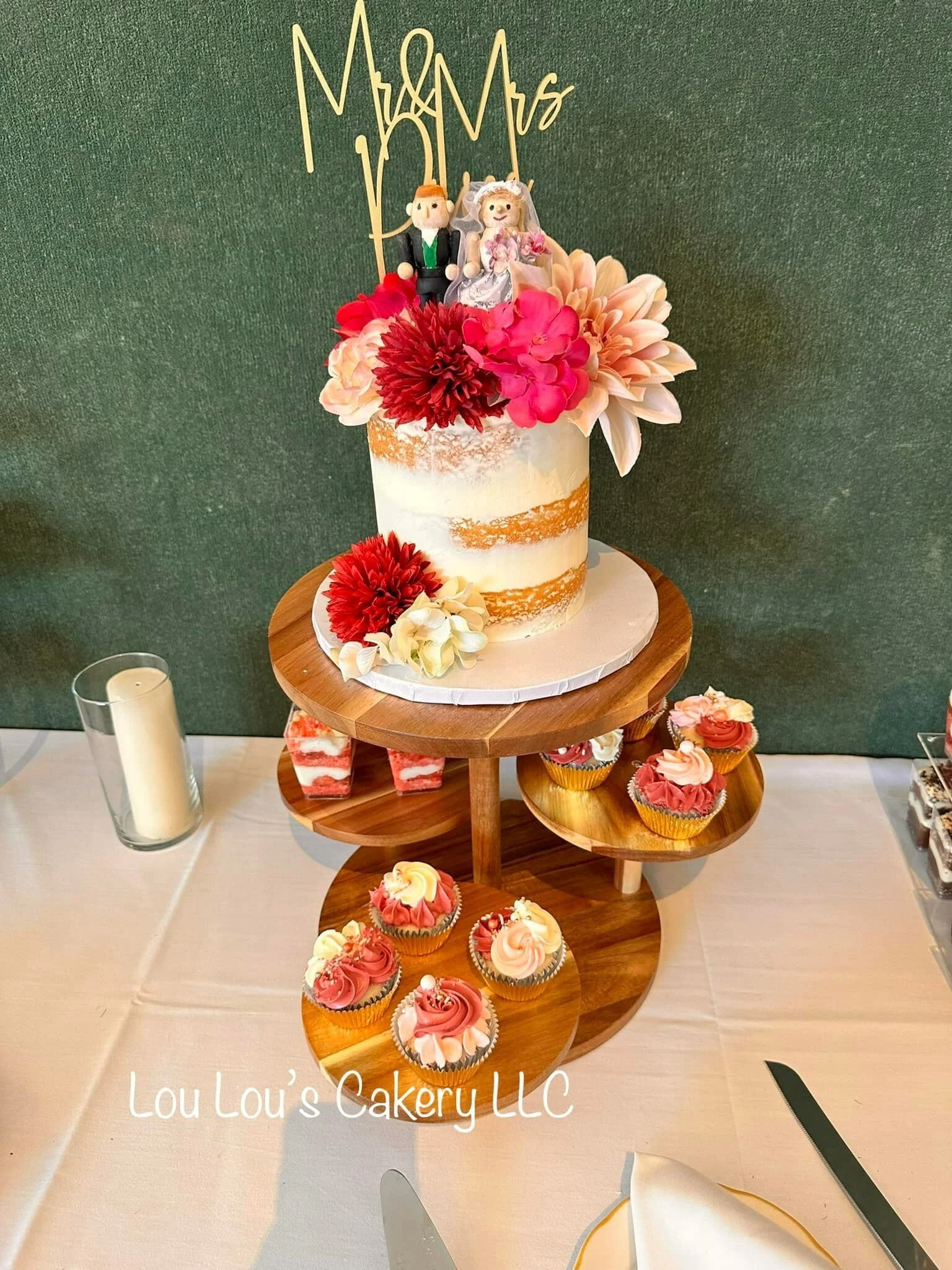 A wedding cake with pink and red flowers, two bride and groom figurines on top, and a 'Mrs. & Mr.' topper, displayed on a wooden stand with cupcakes underneath.