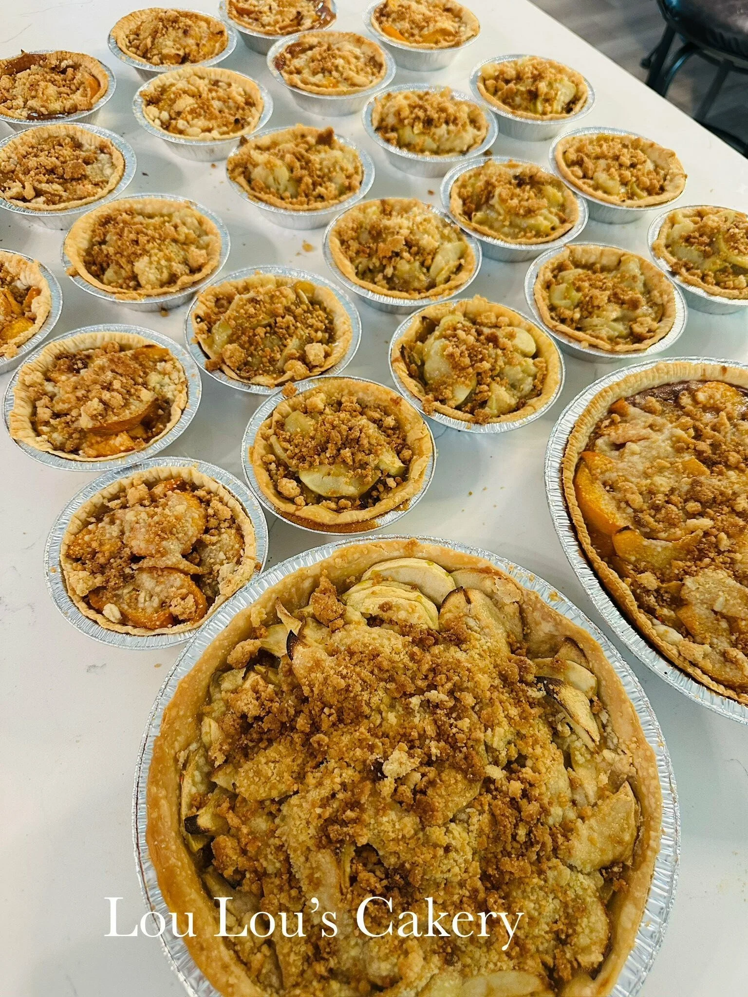 Multiple apple crumb pies in aluminum pans, with a large pie in the foreground and smaller pies in the background, on a white table at Lou Lou's Cakery.