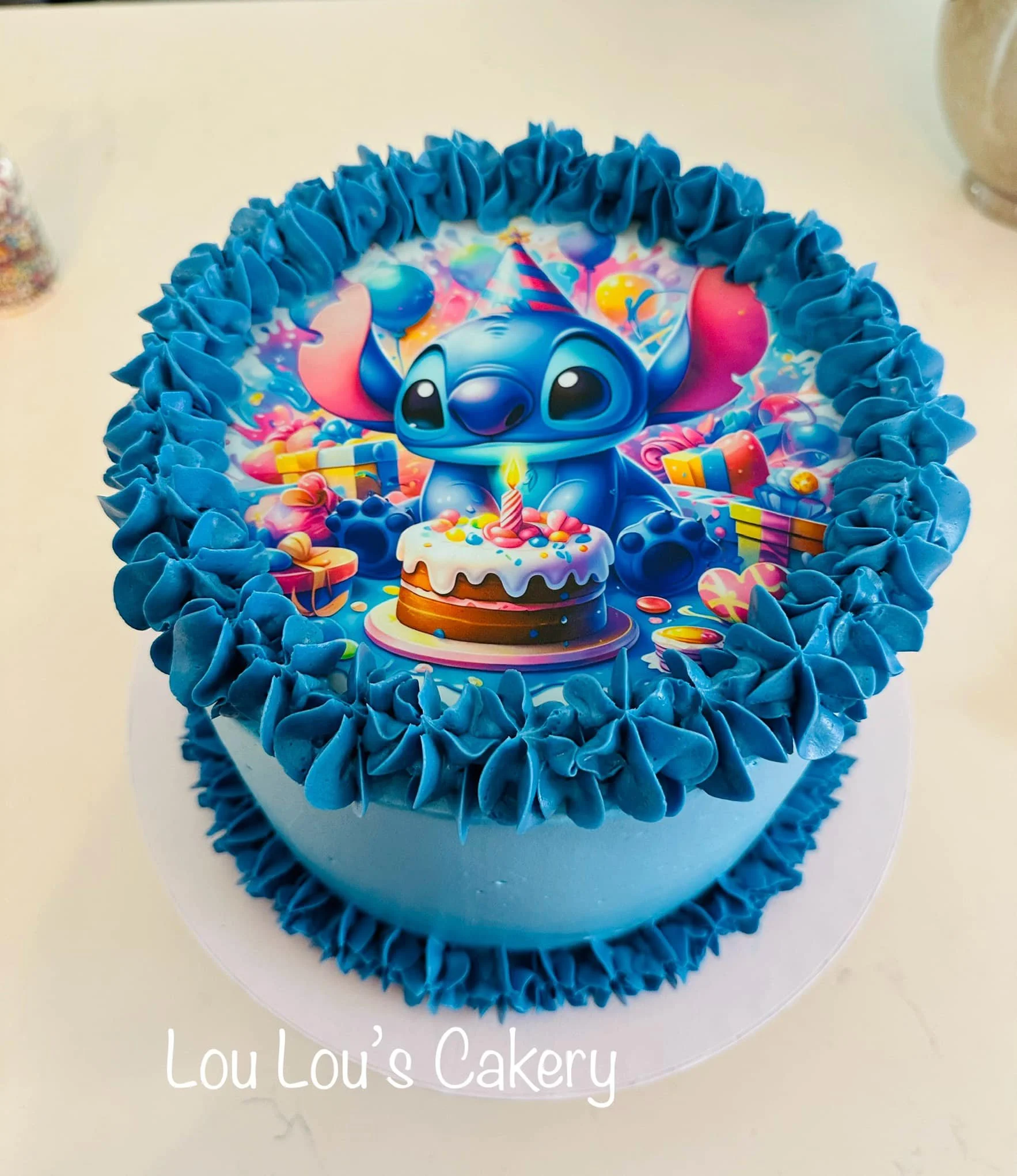 Blue-themed birthday cake with an edible image of Stitch from Lilo & Stitch wearing a party hat, surrounded by colorful balloons, gifts, and a birthday cake graphic.